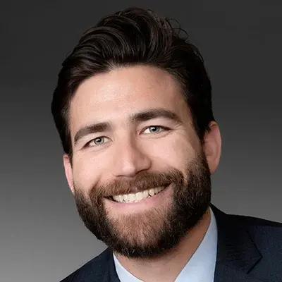 Headshot of a smiling bearded man in a dark suit and light blue shirt against a gray background.