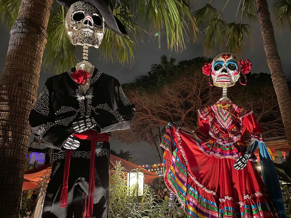 Two tall skeleton figures in a black charro suit and a red folk dress stand under palm trees at night.