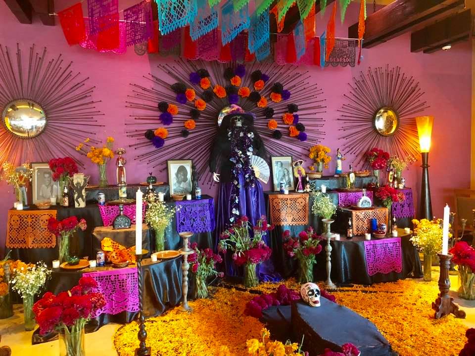 A Day of the Dead altar with marigolds, framed photos, candles, papel picado banners, and a costumed skeleton figure.