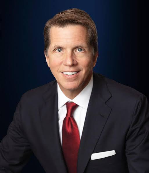 Smiling man in a dark suit, white shirt, and red tie against a dark blue background.