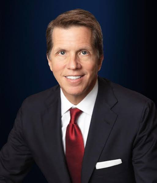 Smiling man in a dark suit, white shirt, and red tie against a dark blue background.
