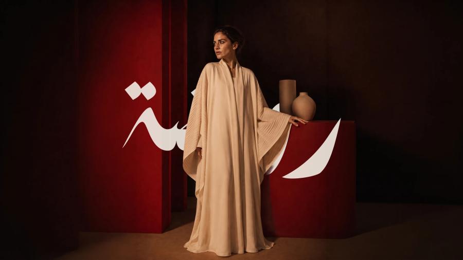 A model in a floor-length camel abaya beside a red plinth of stoneware vessels, the Rawda mark in Arabic behind her.
