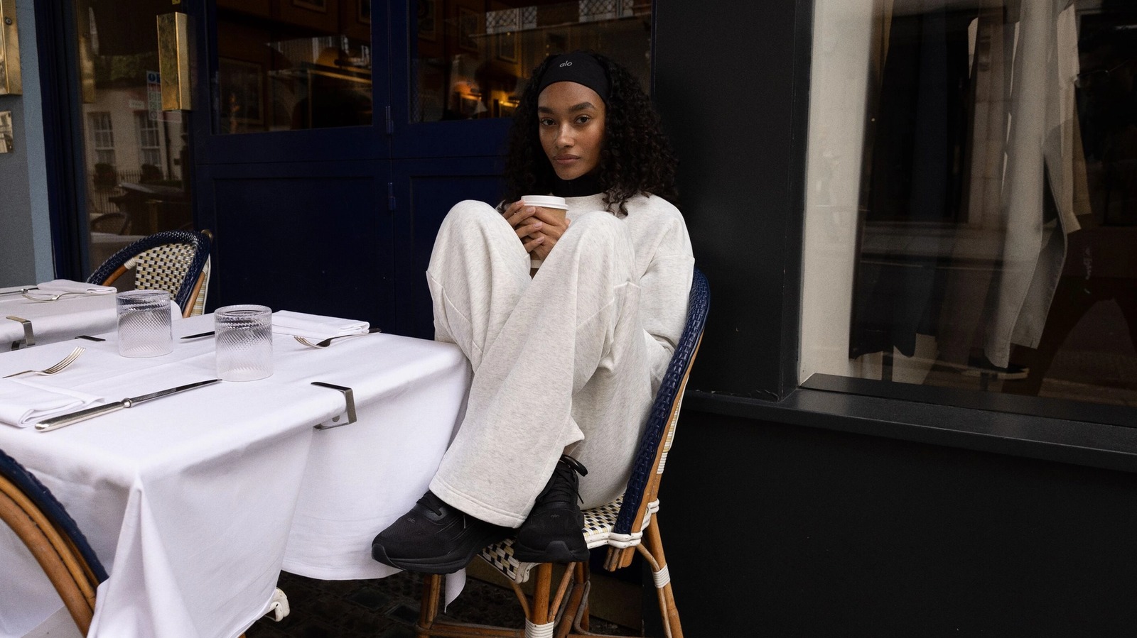 A person in a cozy white outfit sits on a chair outside a cafe, holding a cup. Their expression is calm and introspective. Tables are set with white cloths, glassware, and cutlery.