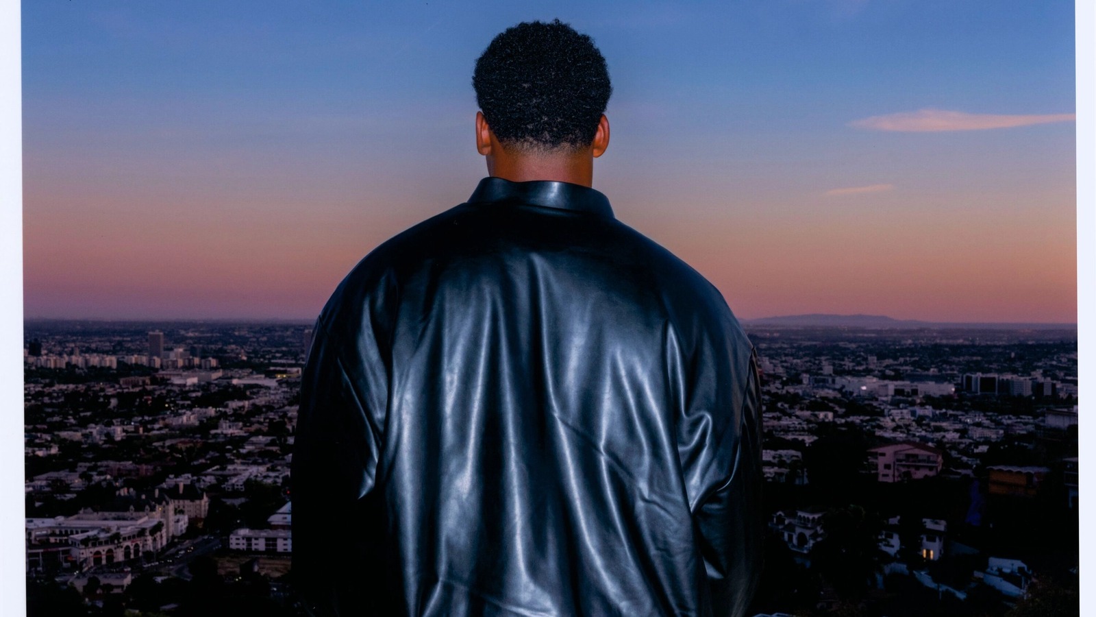 A person in a black leather jacket stands on a balcony overlooking a city at sunset. The sky is a gradient of pink and blue, creating a serene mood.