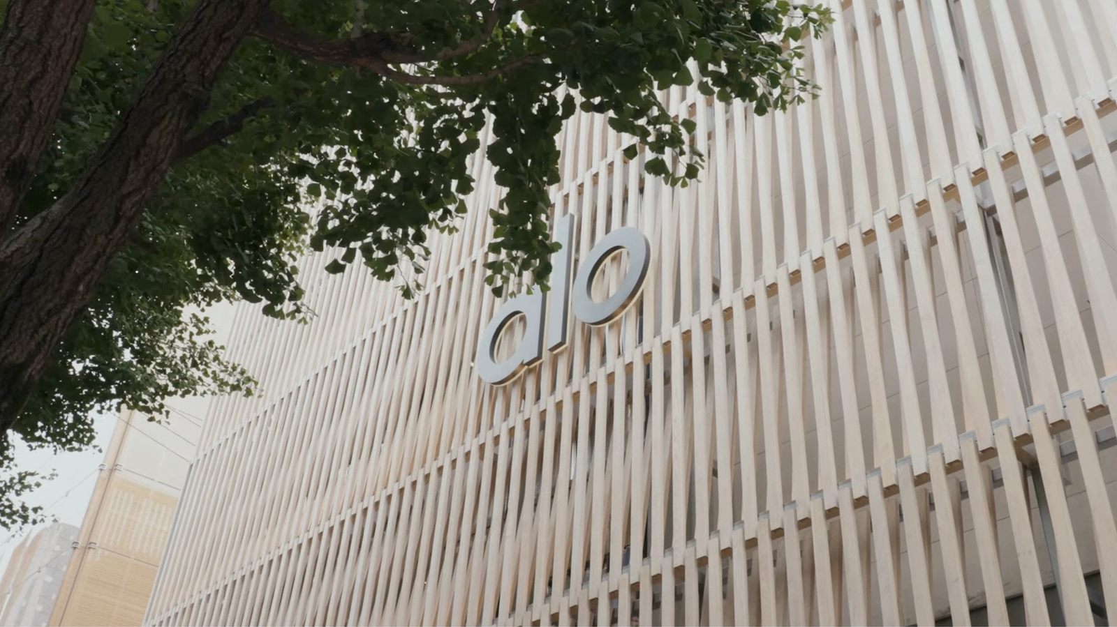 A beige building facade with vertical slats and the letters "do" prominently displayed. A leafy tree branches over the structure, creating a serene tone.