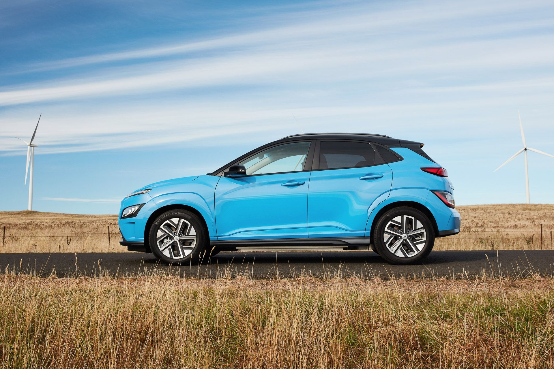 Blue with black roof Hyundai Kona Electric side view in front of wind turbines