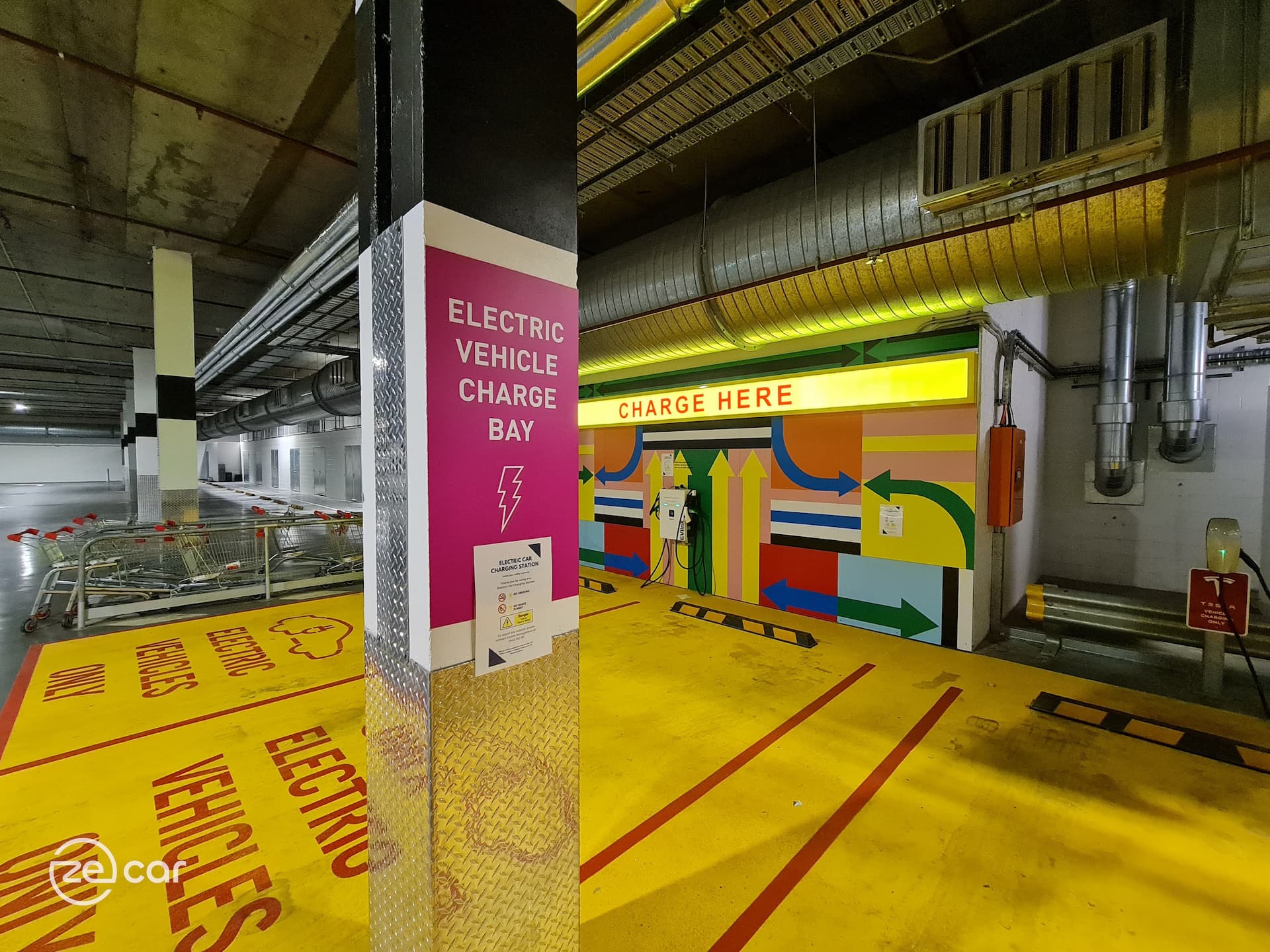 EV AC charger with yellow 'Charge Here' sign and pink 'Electric Vehicle Charge Bay' sign