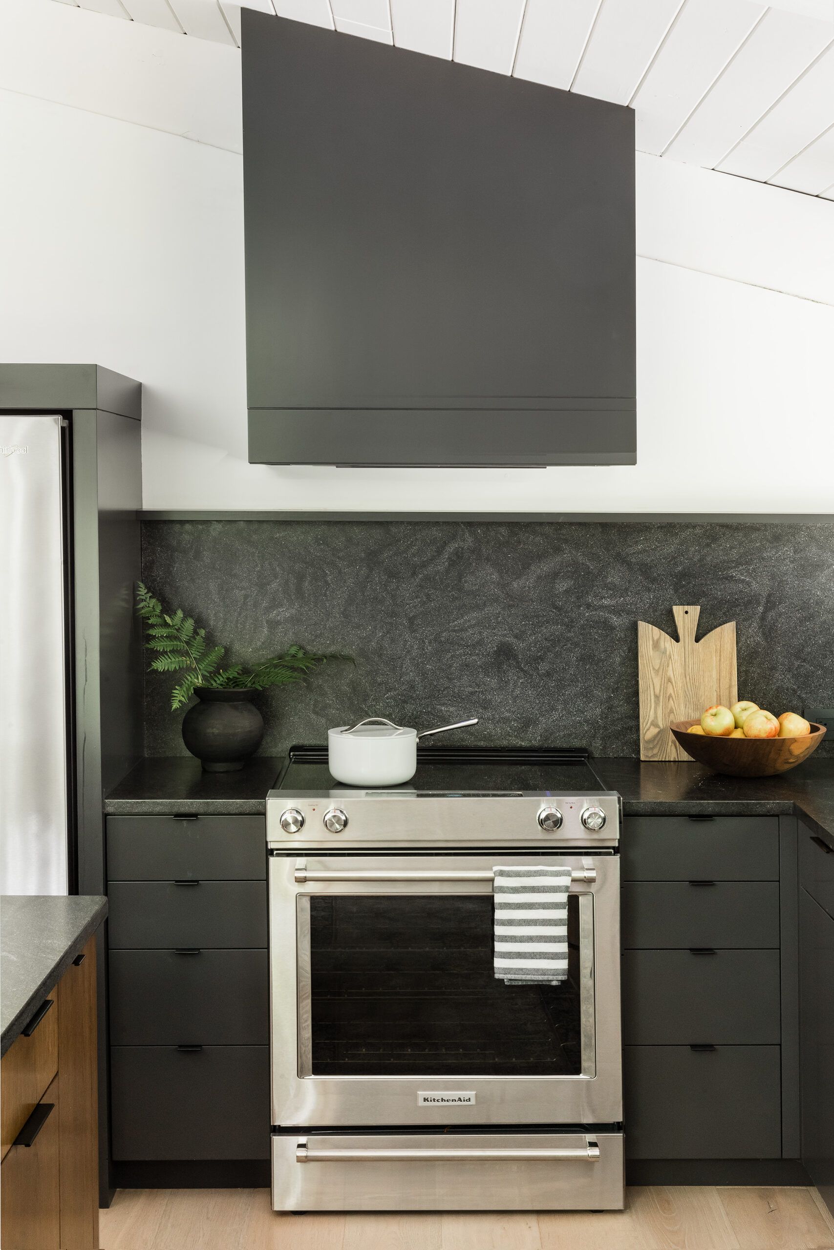 A stainless steel range under a charcoal range hood, flanked by dark stone counters, a cutting board, and a bowl of apples.