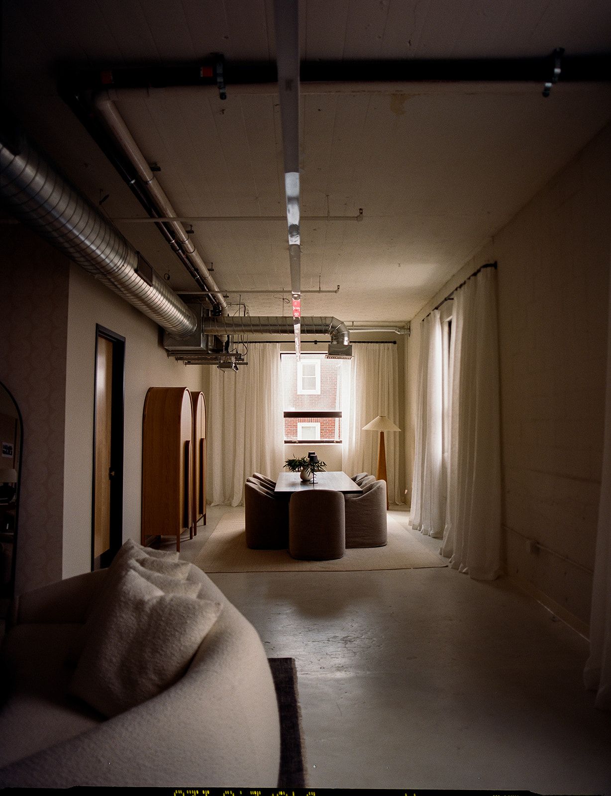 Dimly lit dining area with ductwork overhead, a dark wood table, upholstered chairs, sheer curtains, and a tall wood floor lamp.
