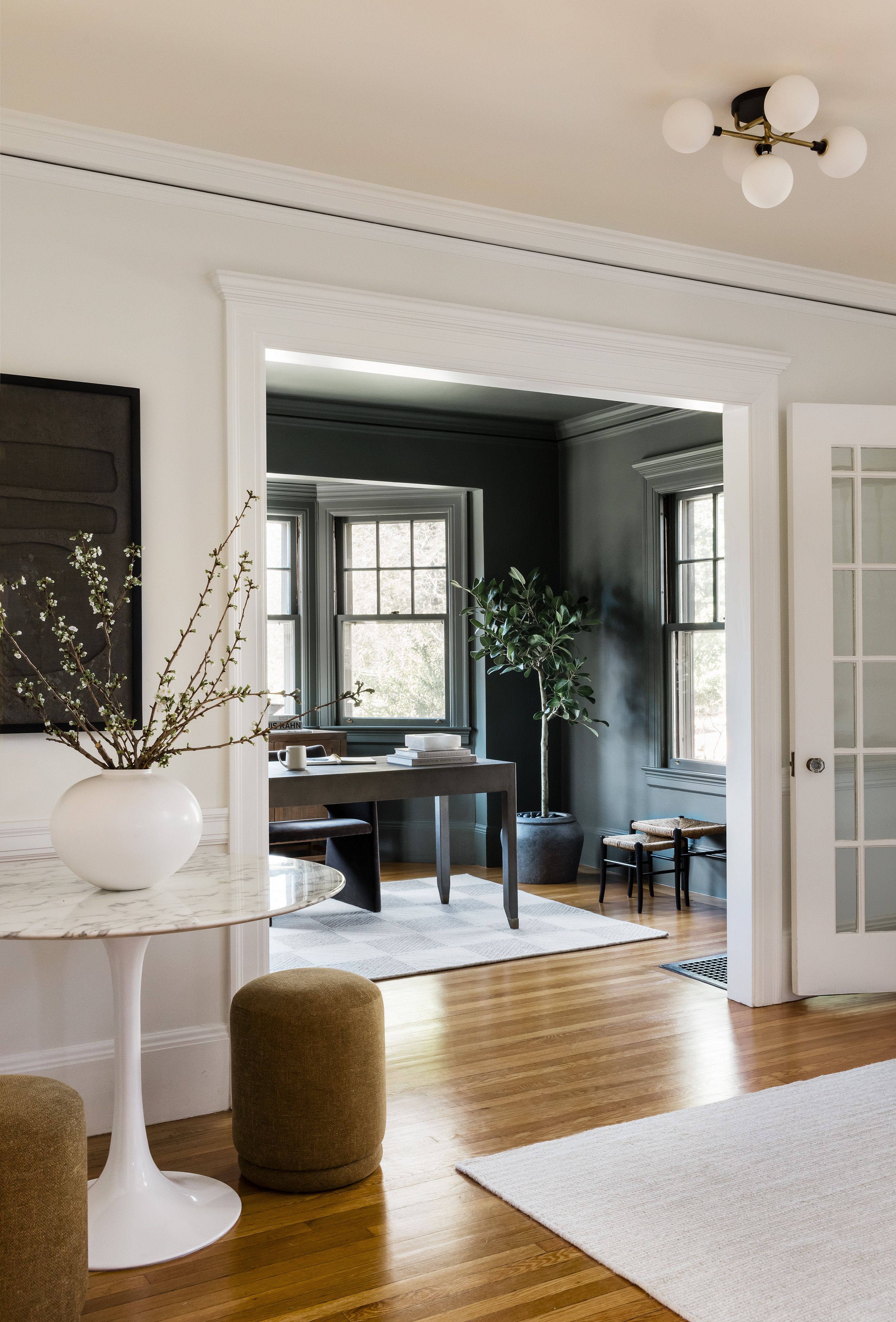 An entryway with a round marble table and branches in a white vase, opening to a dark green home office with a wood desk.