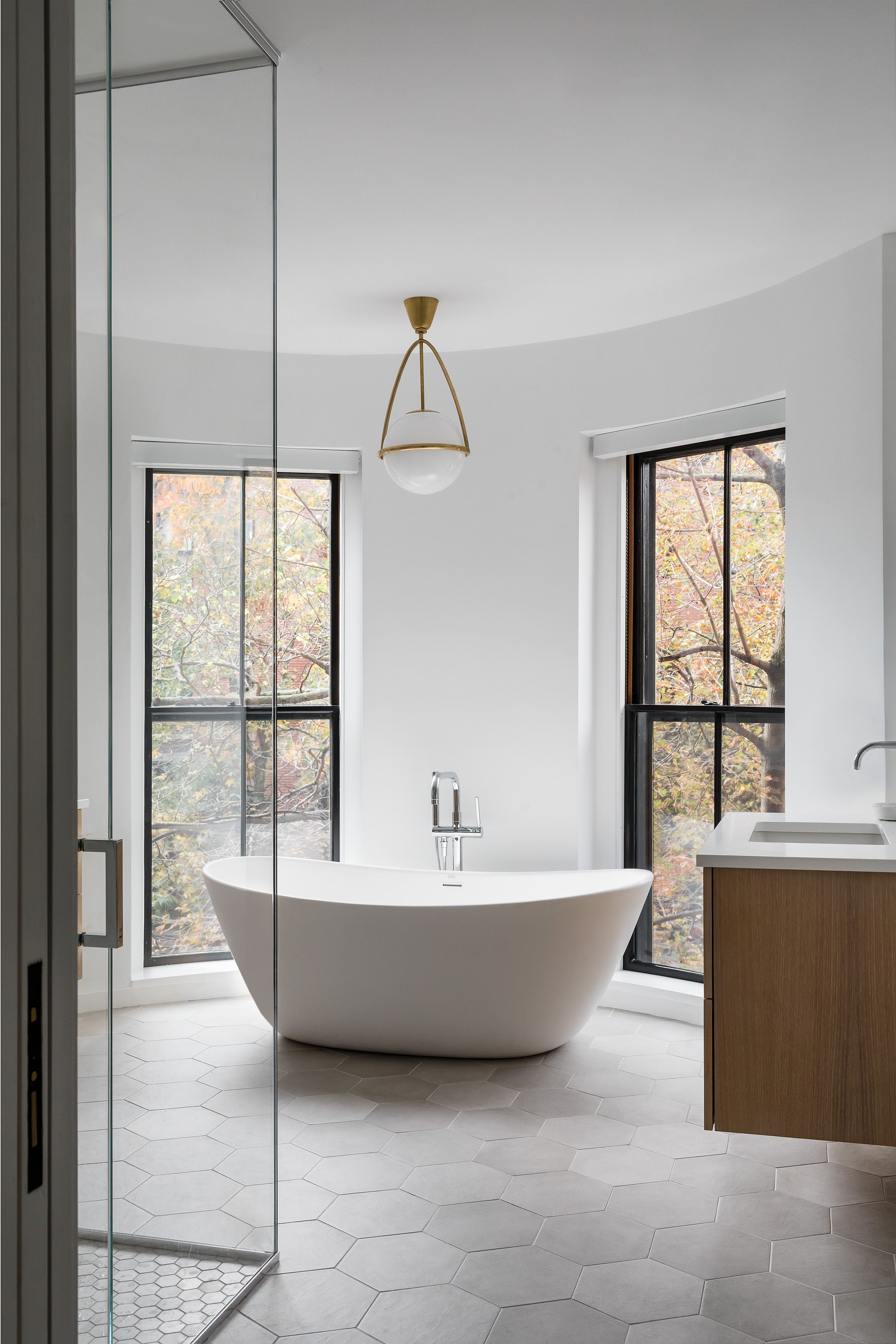A primary bathroom with a curved bay window, freestanding oval tub, brass pendant light, and gray hexagon tile floor.