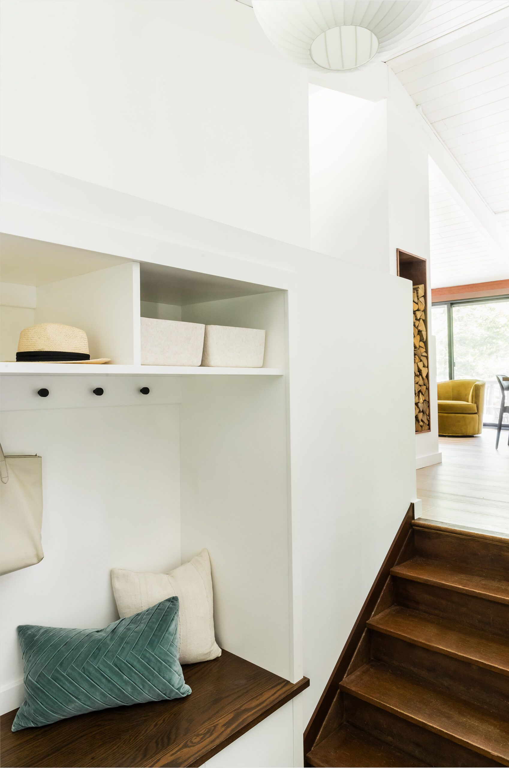 A mudroom nook with a built-in bench, open cubbies holding a straw hat, and teal and linen pillows beside wood stairs.