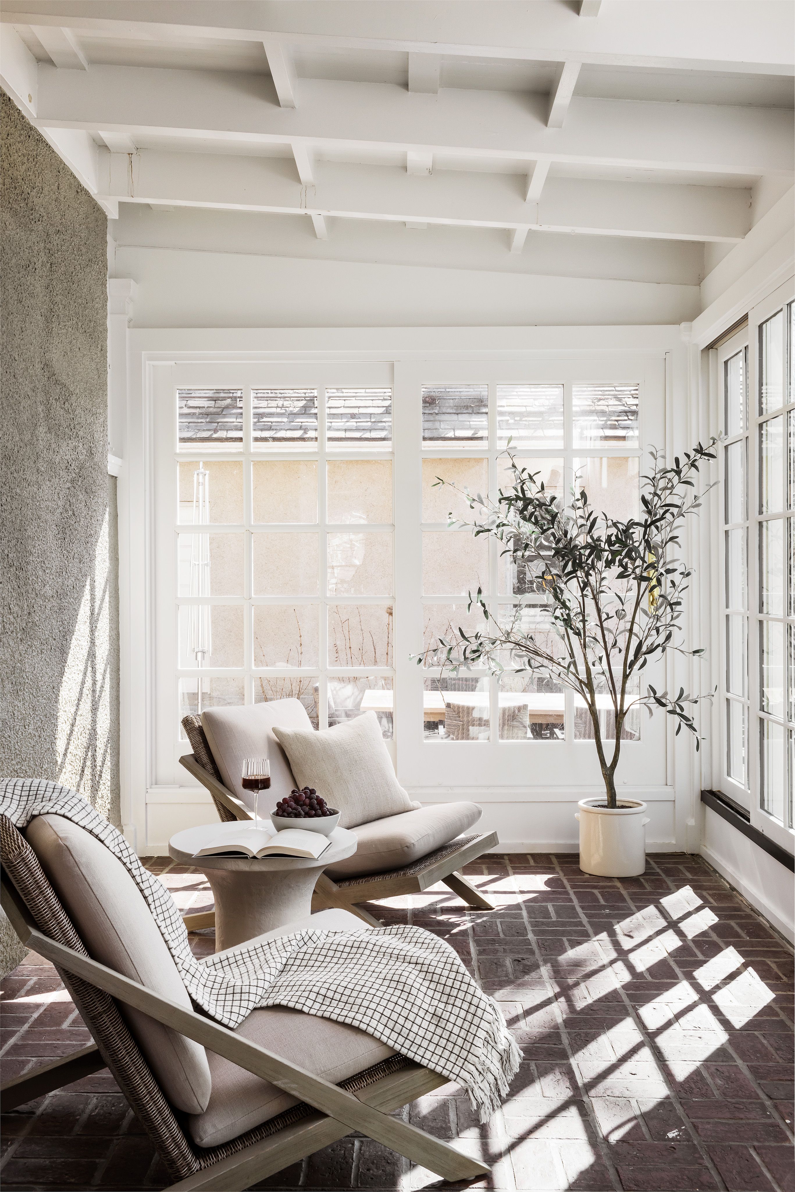 A sunroom with a brick floor, woven lounge chairs draped in a checked throw, a potted olive tree, and white beamed ceilings.