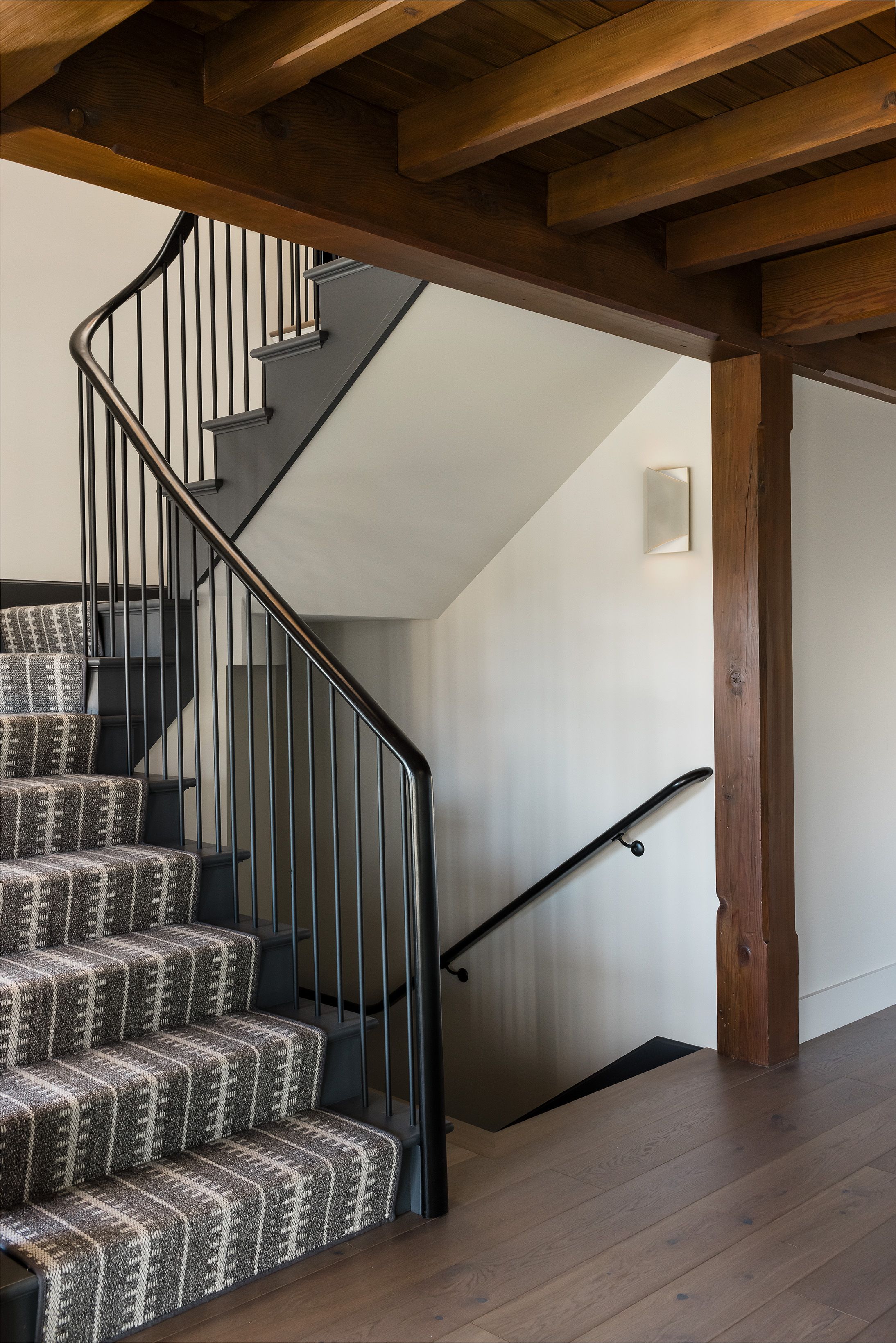 A wood-beamed stairwell has a curved black metal railing and a patterned charcoal-and-cream stair runner leading to wide-plank floors.