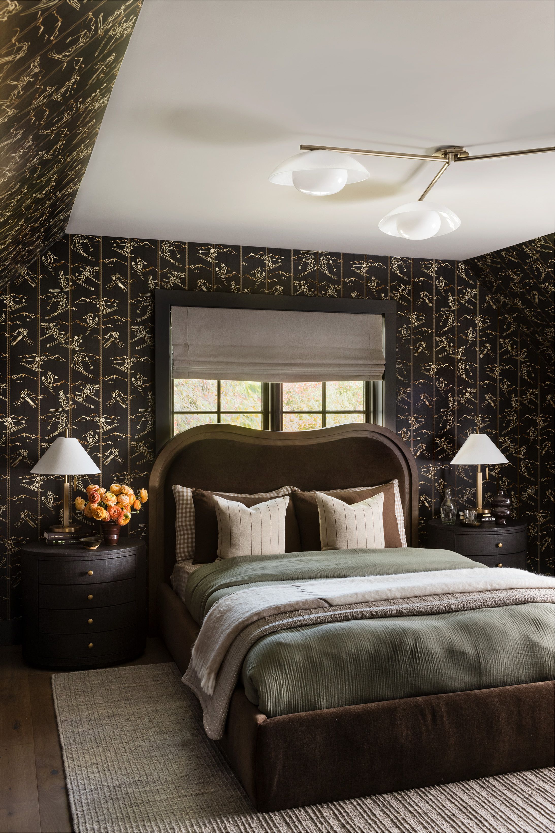 A dark bedroom with gold ski-print wallpaper pairs a curved brown leather headboard with a built-in bunk alcove and round black nightstands.