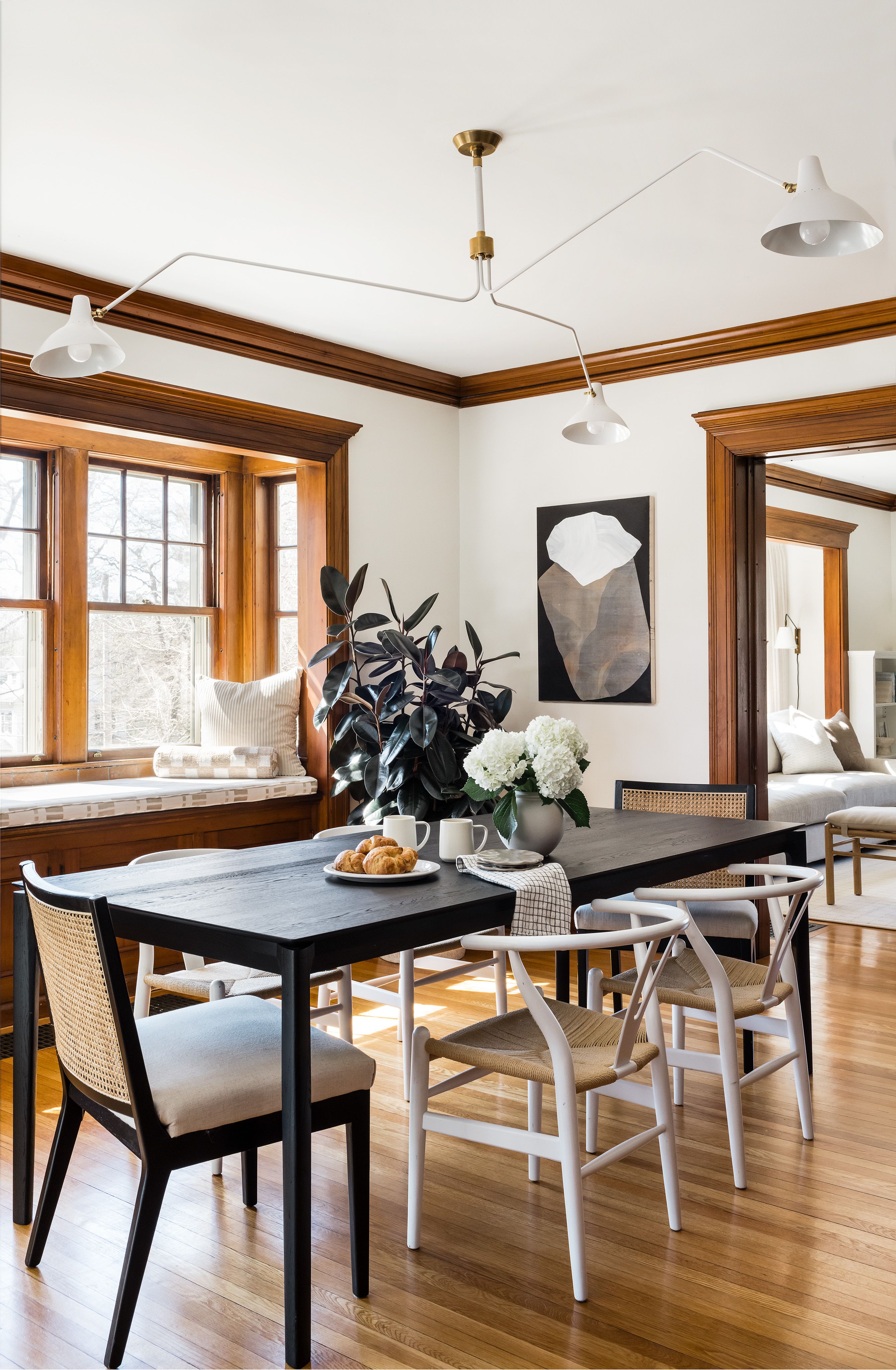 A dining room with a black table, white and black wishbone chairs, a cane-back chair, and abstract art above a built-in window seat.