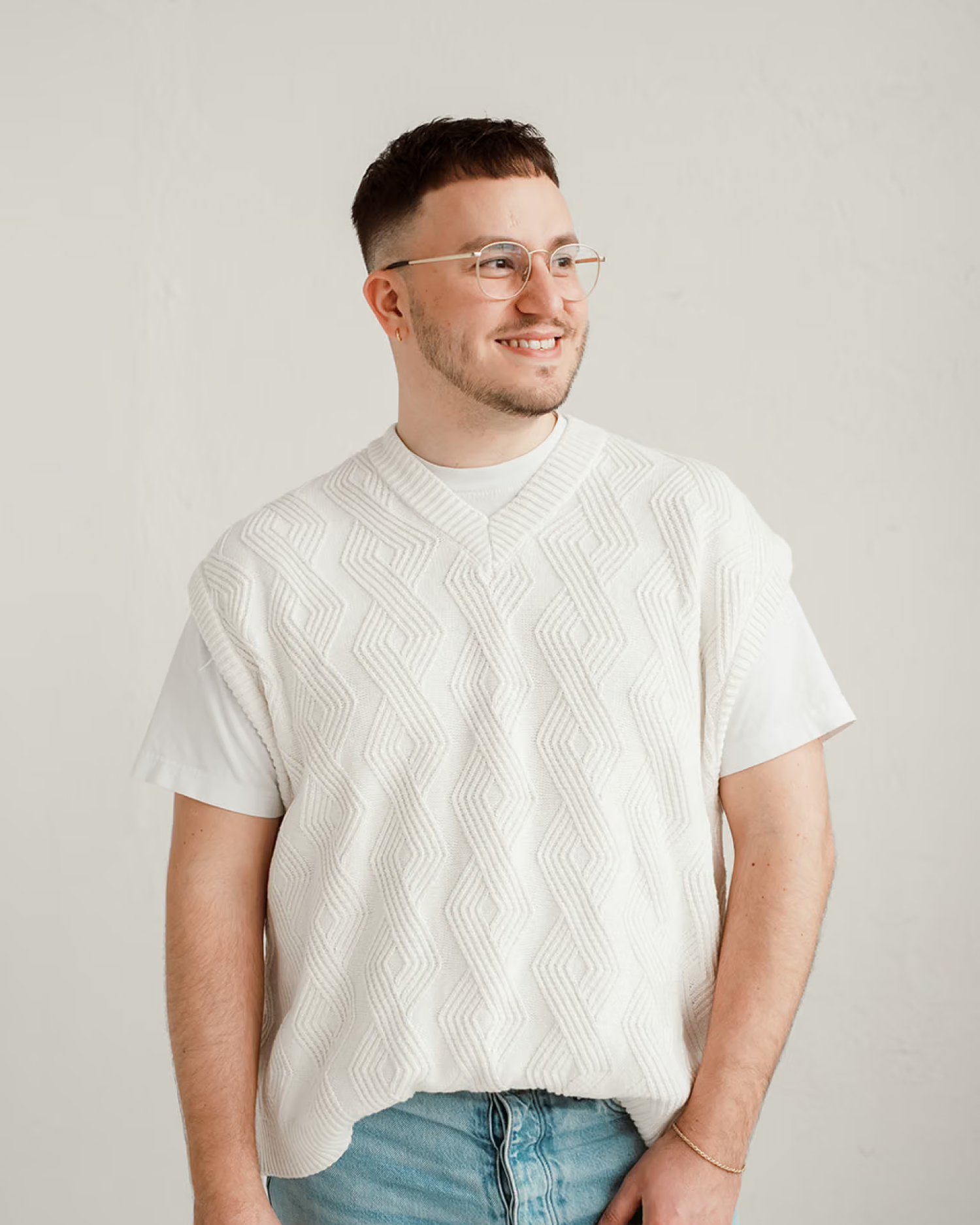 Tyler Poquette smiles wearing a textured ivory sweater vest over a white T-shirt against a plain backdrop.