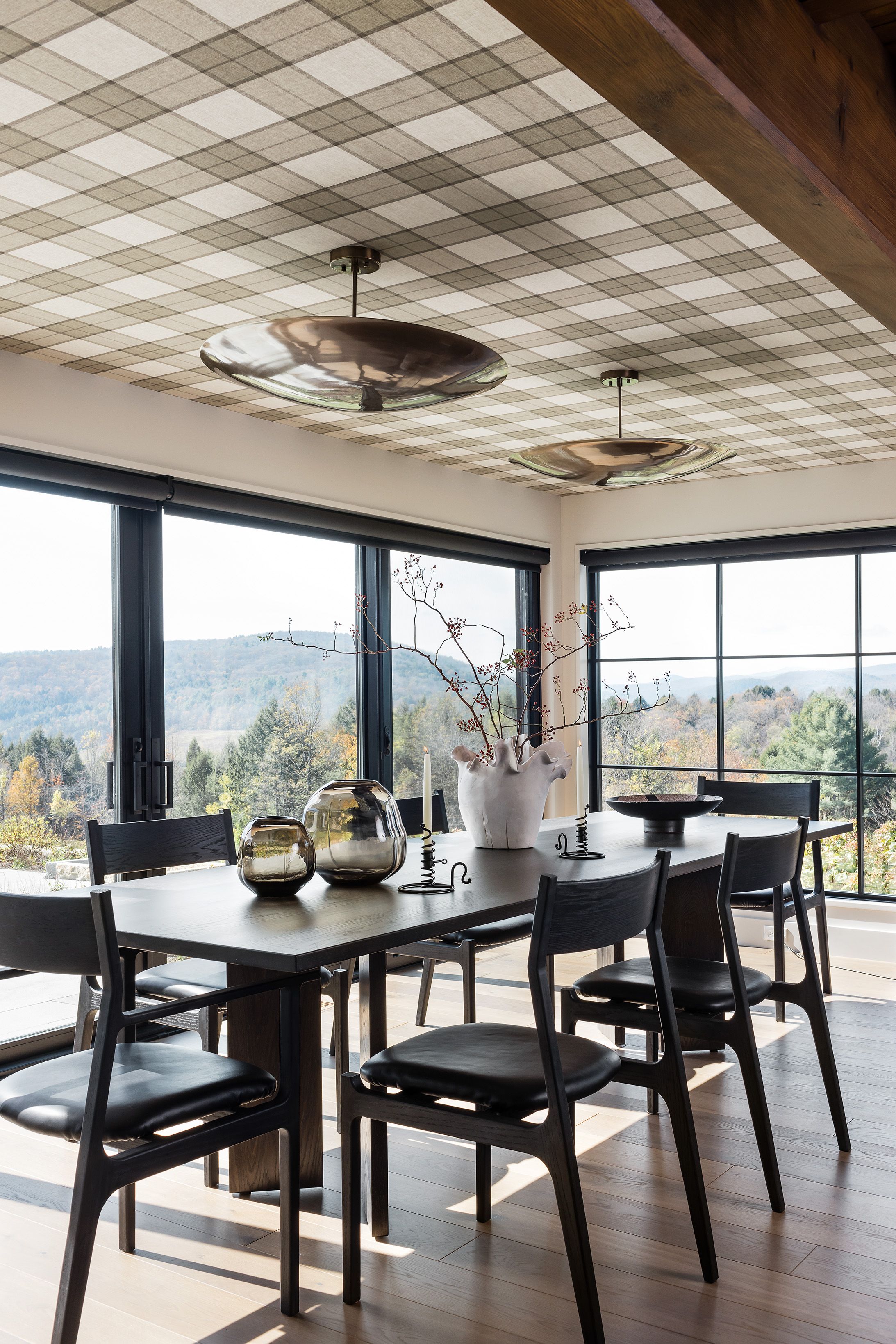 A dining room with a plaid-papered ceiling and two brass disc pendants sits beside floor-to-ceiling windows framing a mountain view.