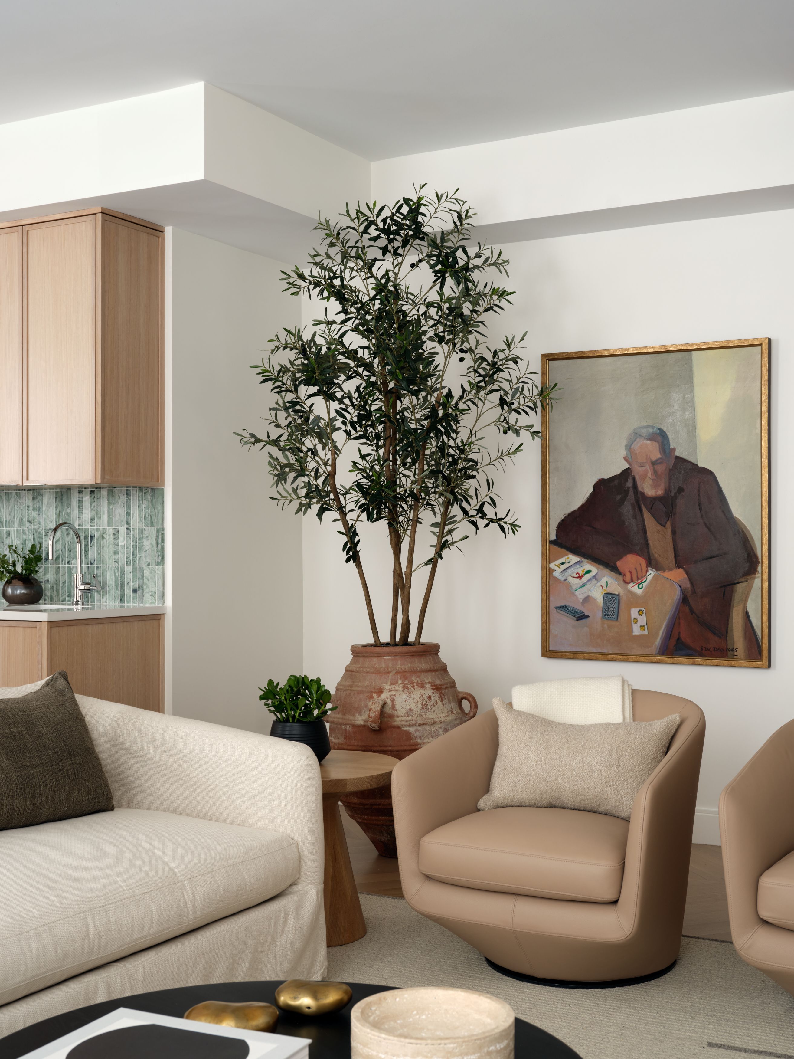 A living room corner with a potted olive tree in a terracotta urn, a gold-framed portrait painting, and tan leather swivel chairs.
