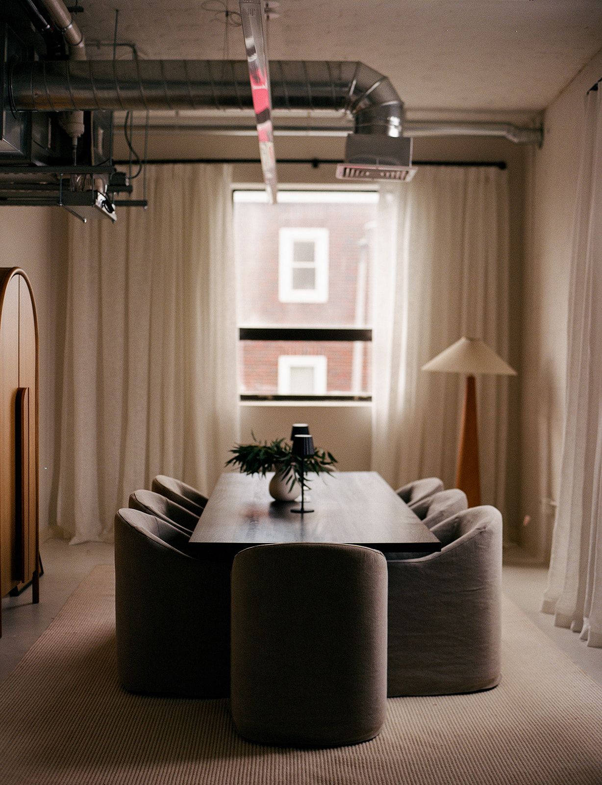 Dining area with a dark wood table, six curved taupe barrel chairs, a wood floor lamp with an angled shade, and sheer curtains.