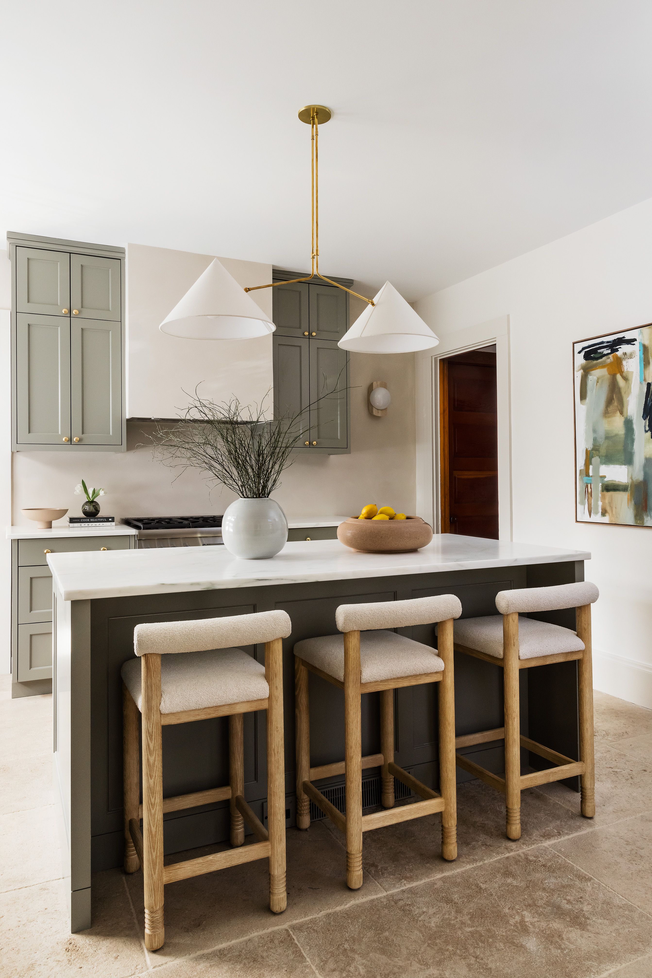 A kitchen island with a brass branching pendant light, boucle-seated wood stools, and a view into a wood-paneled butler's pantry.
