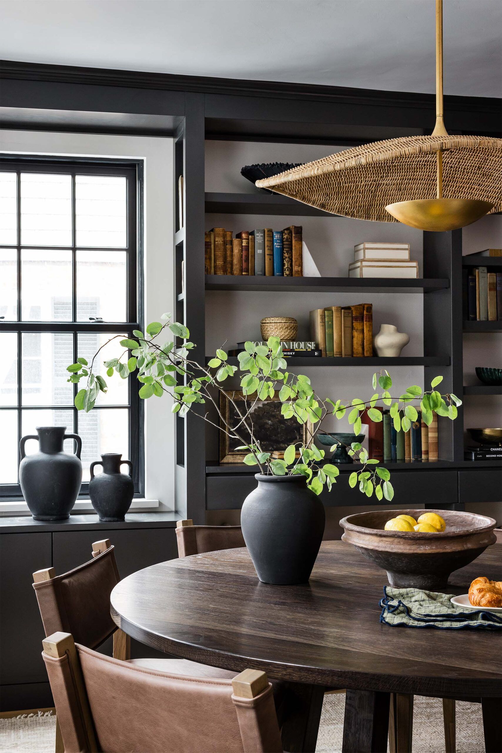 Dark dining area with black built-in bookshelves, a woven rattan and brass pendant light, and a round wood table set with lemons.