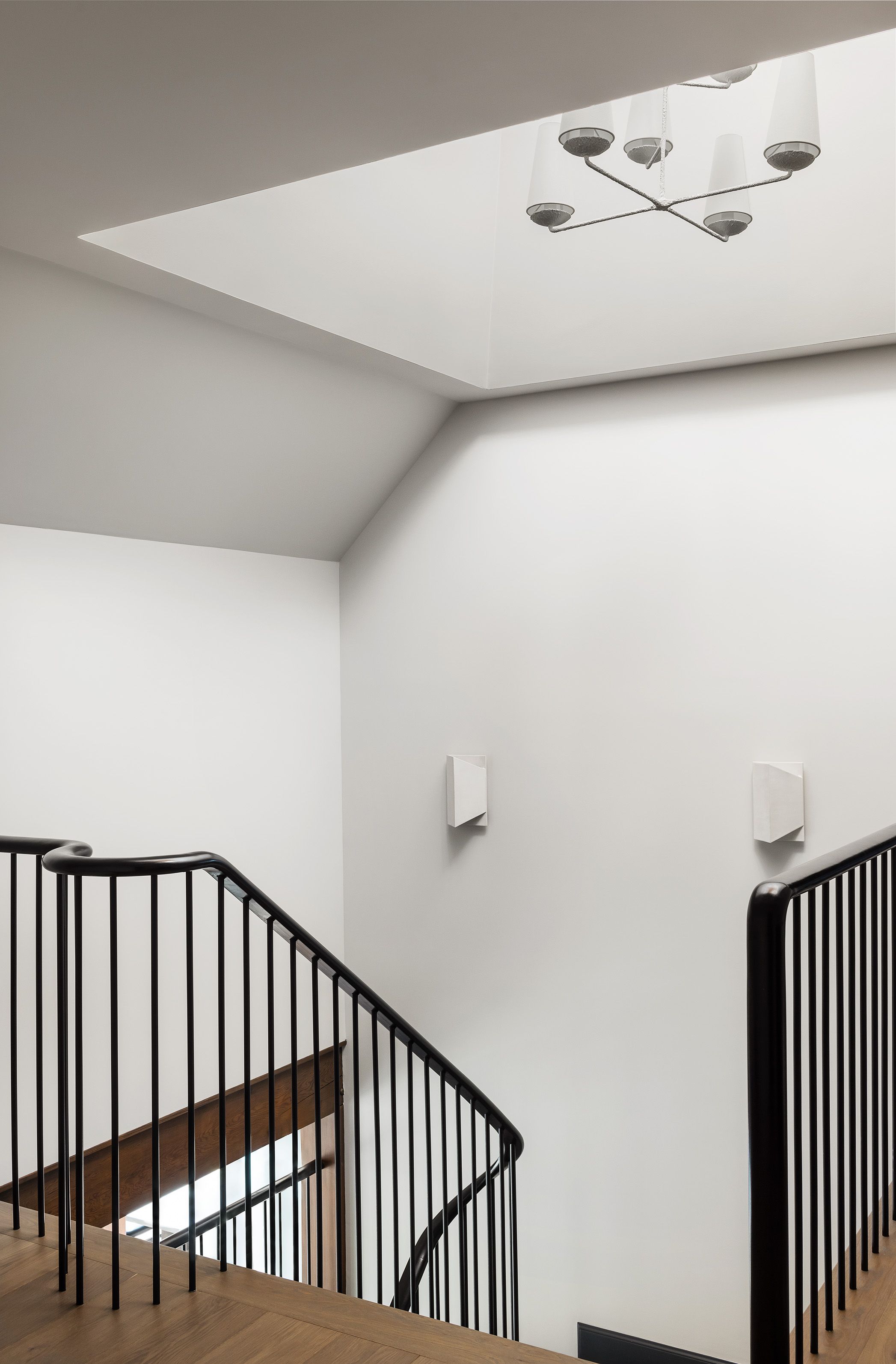 A stairwell lit by an overhead skylight has a sculptural white ceiling fixture and black metal railings framing the steps below.