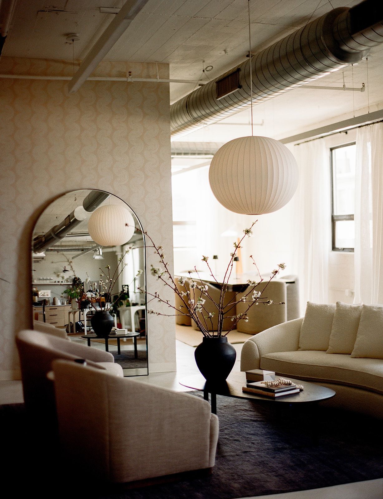 A loft lounge with a curved ivory sofa, flowering branches in a black vase, round paper pendant lights, and patterned wallpaper.