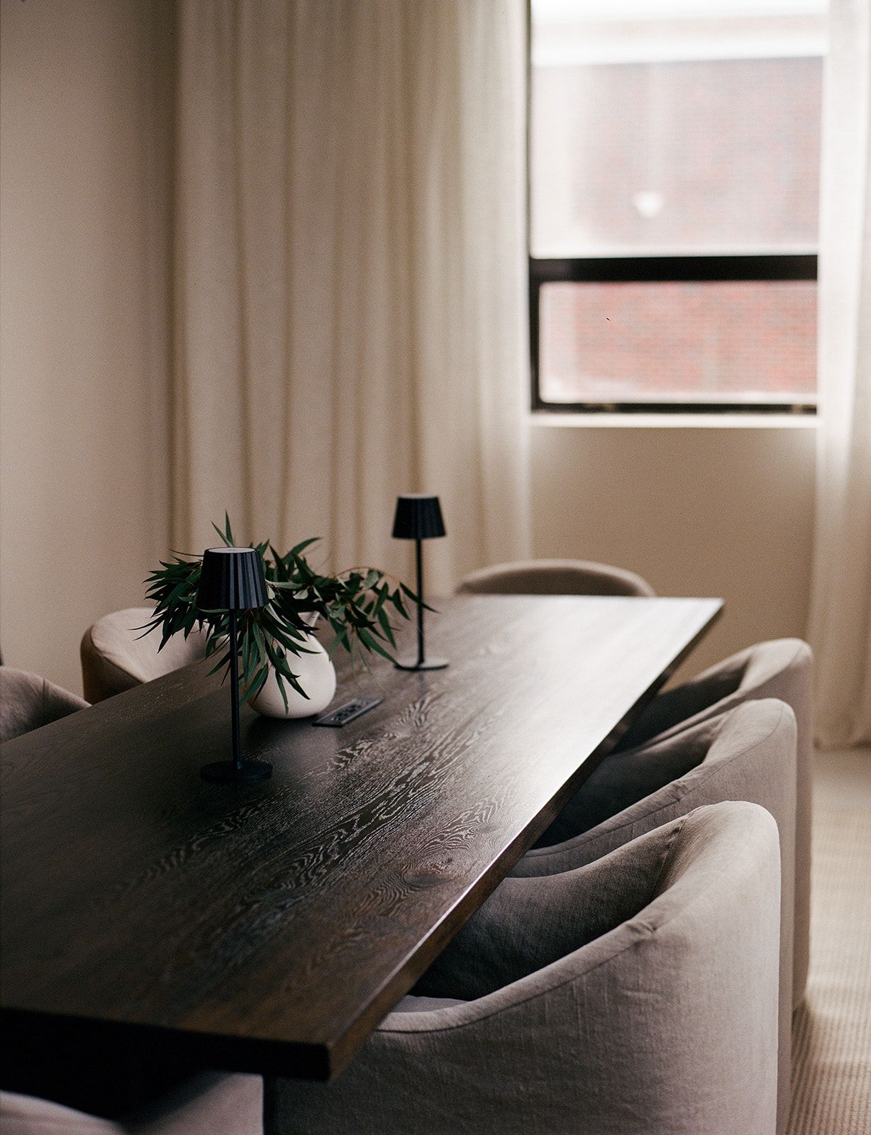 Close-up of a dark wood dining table holding slim black table lamps and a sprig of eucalyptus, with beige chairs blurred behind.