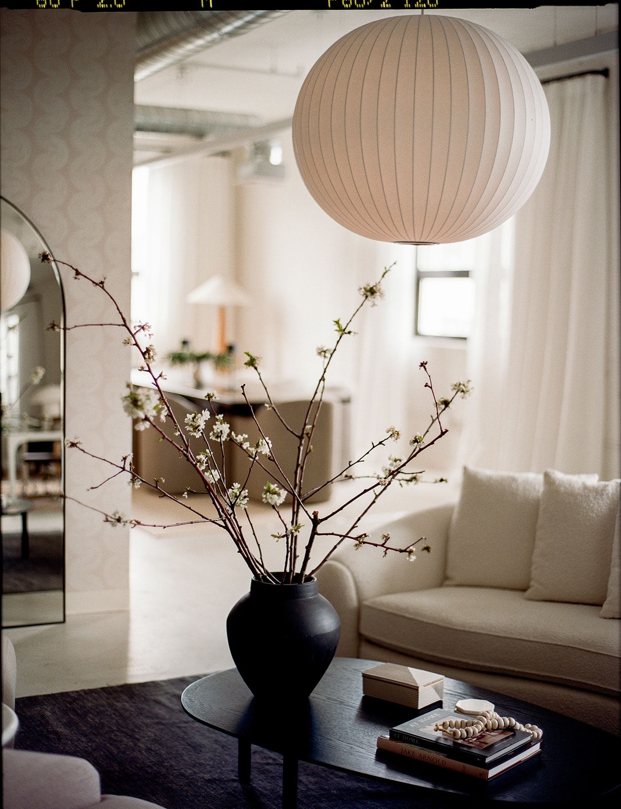 Flowering branches in a black ceramic vase sit on a black coffee table beneath a round paper lantern pendant near a cream sofa.