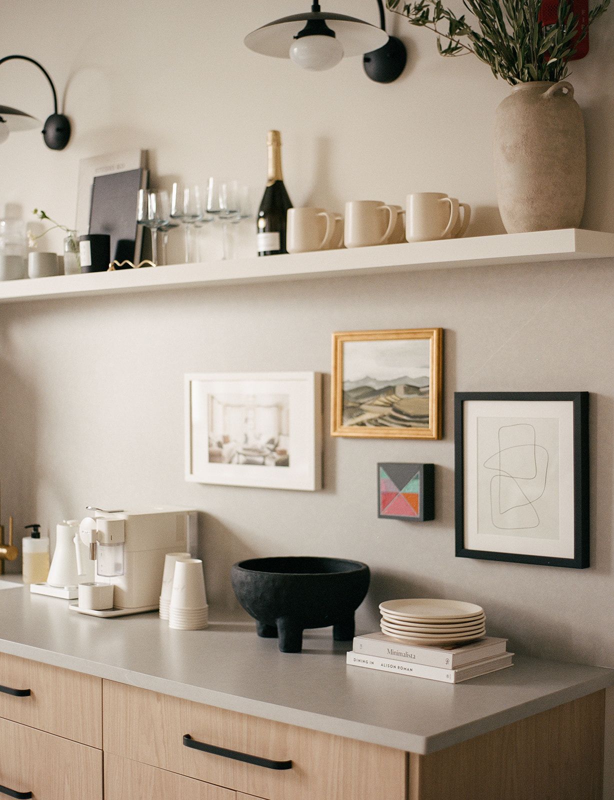 A studio kitchenette shelf with cream mugs, a champagne bottle, wine glasses, and a gallery wall of framed art above stacked plates.