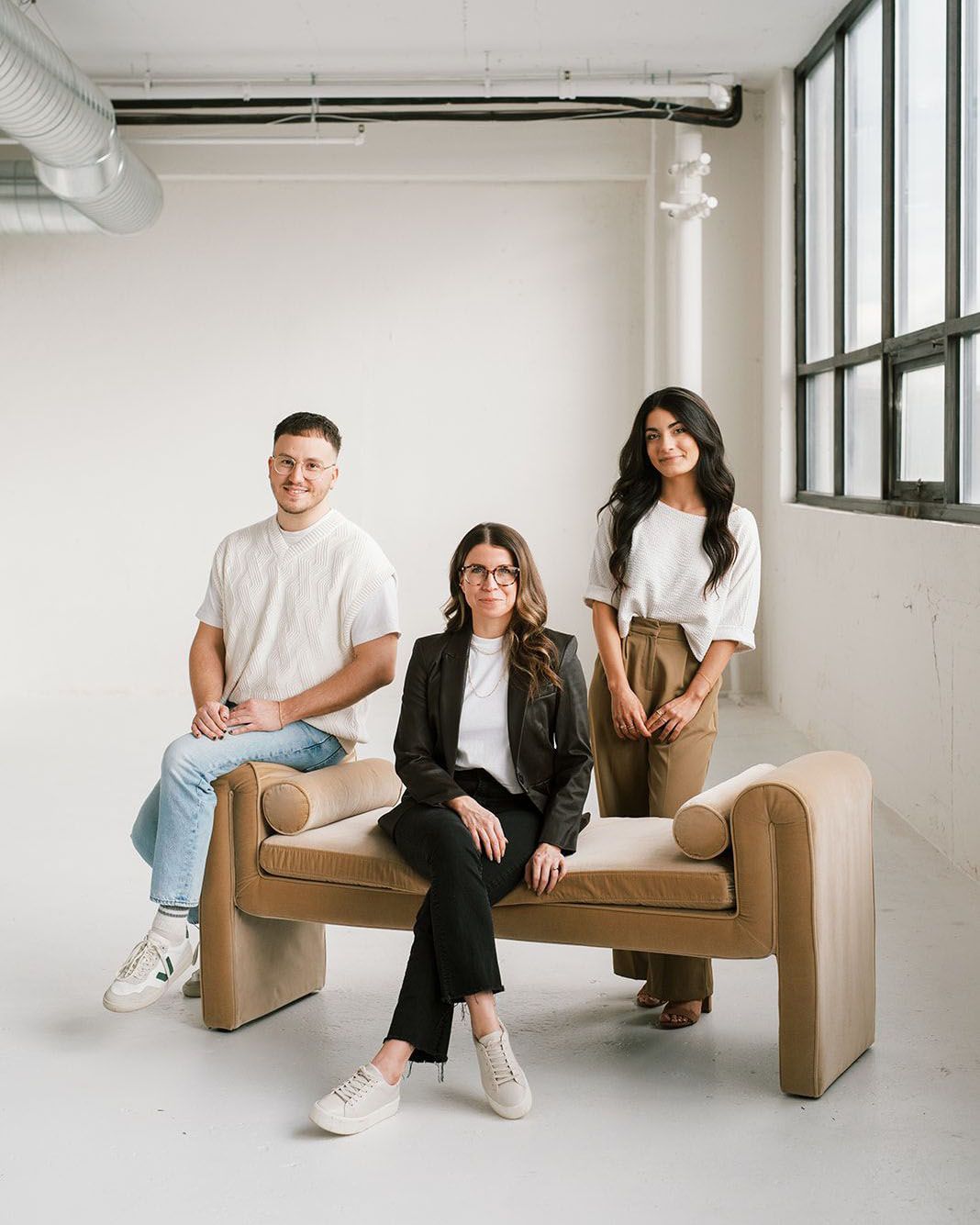 Three team members sit and stand around a camel velvet bench with bolster arms in a bright, industrial studio.