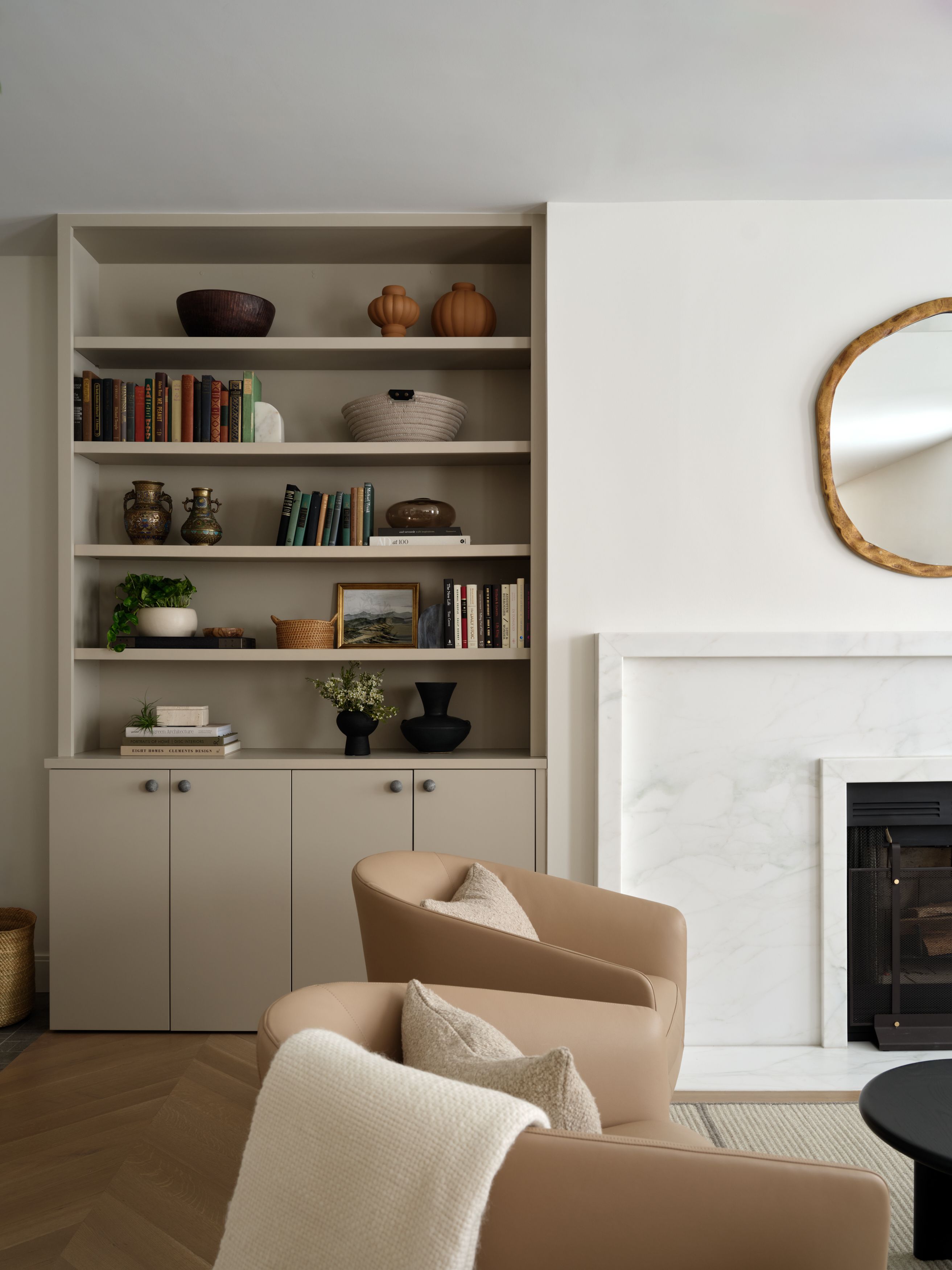 A living room built-in bookshelf styled with ceramics and books beside a marble fireplace and an organic brass mirror.
