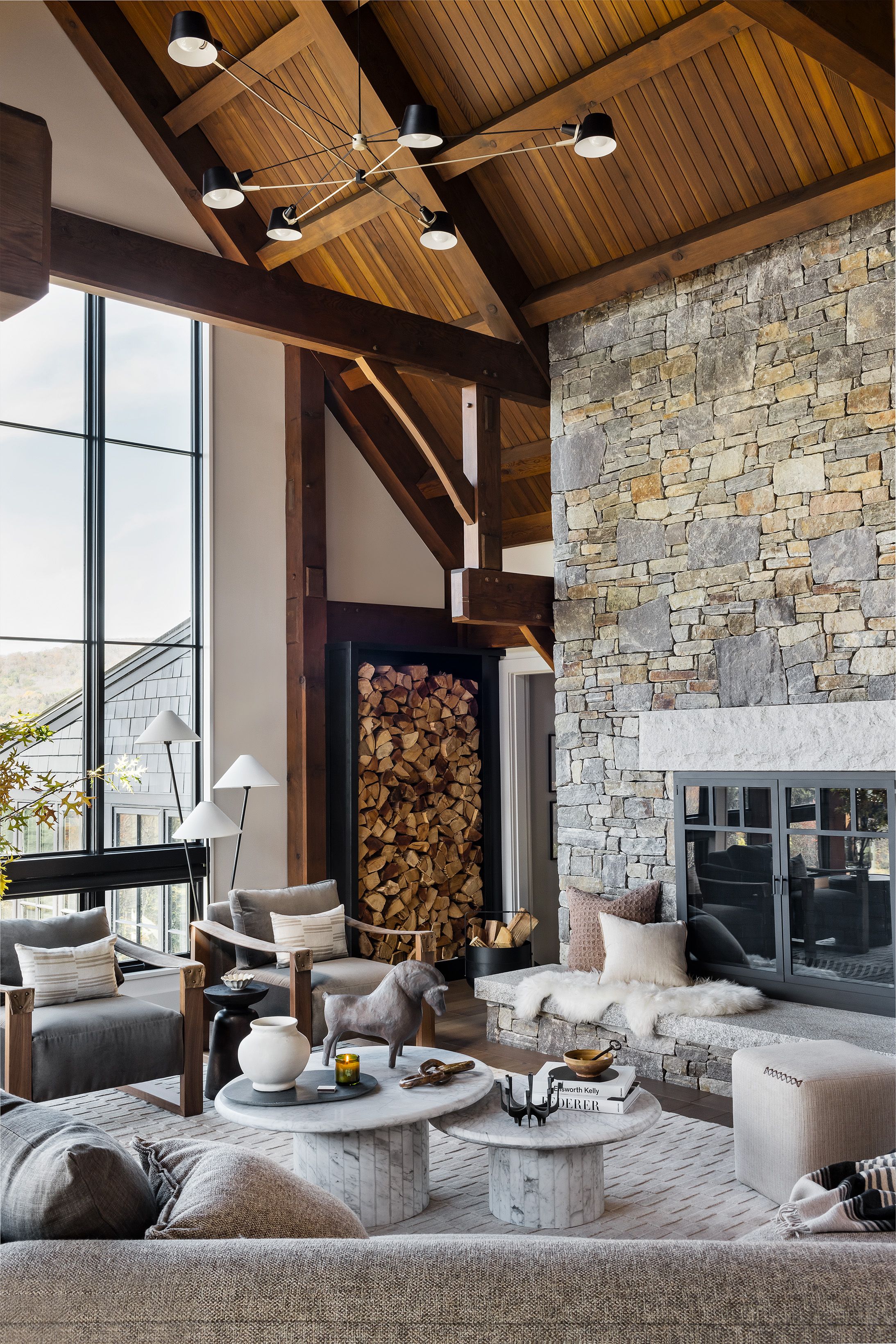 A living room's floor-to-ceiling stone fireplace has a built-in firewood niche, flanked by a timber-beamed ceiling and gray lounge chairs.