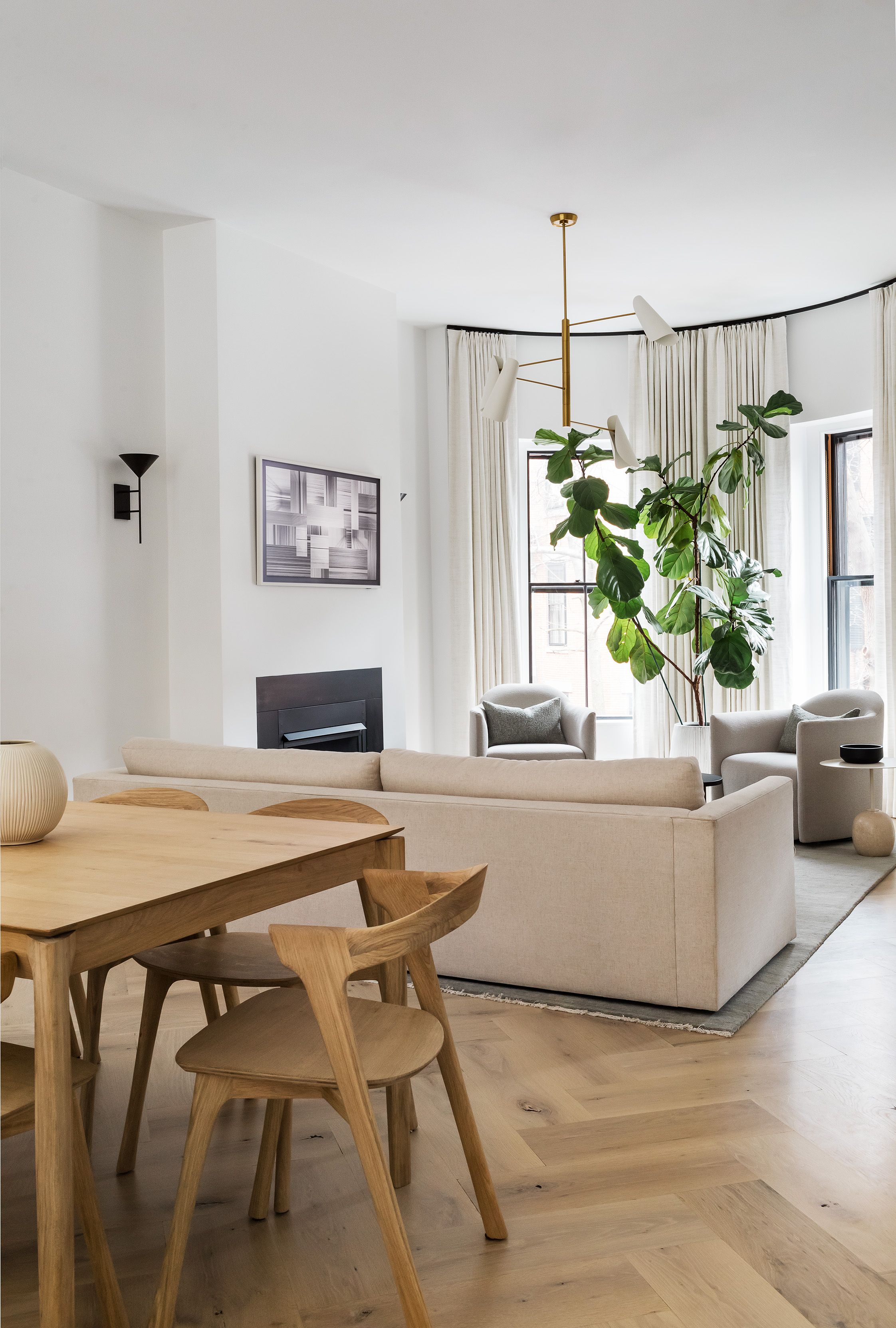 Living and dining space with a wood table, sculptural oak chairs, a cream sofa, a black fireplace, and a fiddle-leaf fig tree.