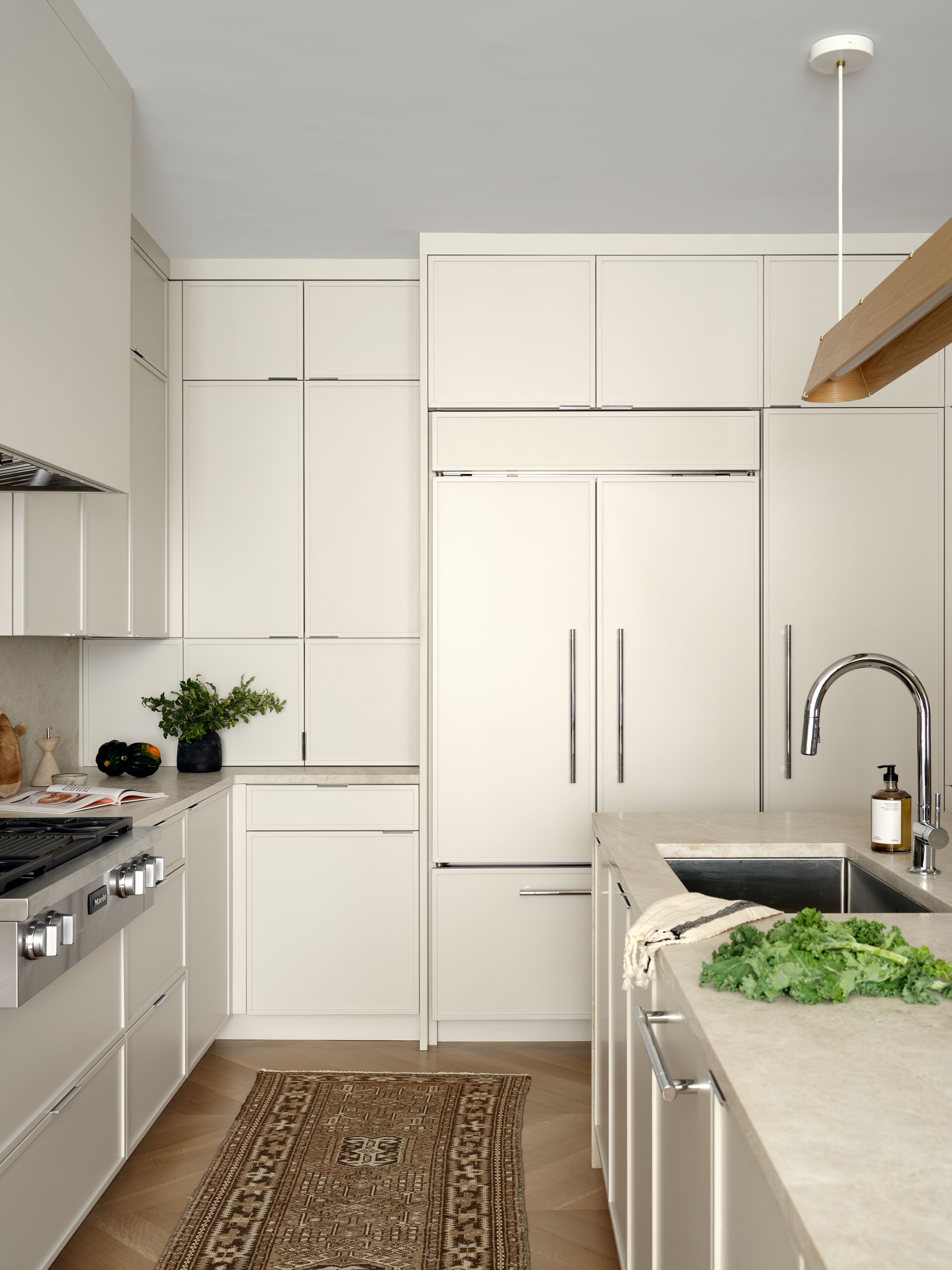 A kitchen with tall cream cabinetry, a paneled refrigerator, a brass pendant light, and a patterned runner on herringbone floor.