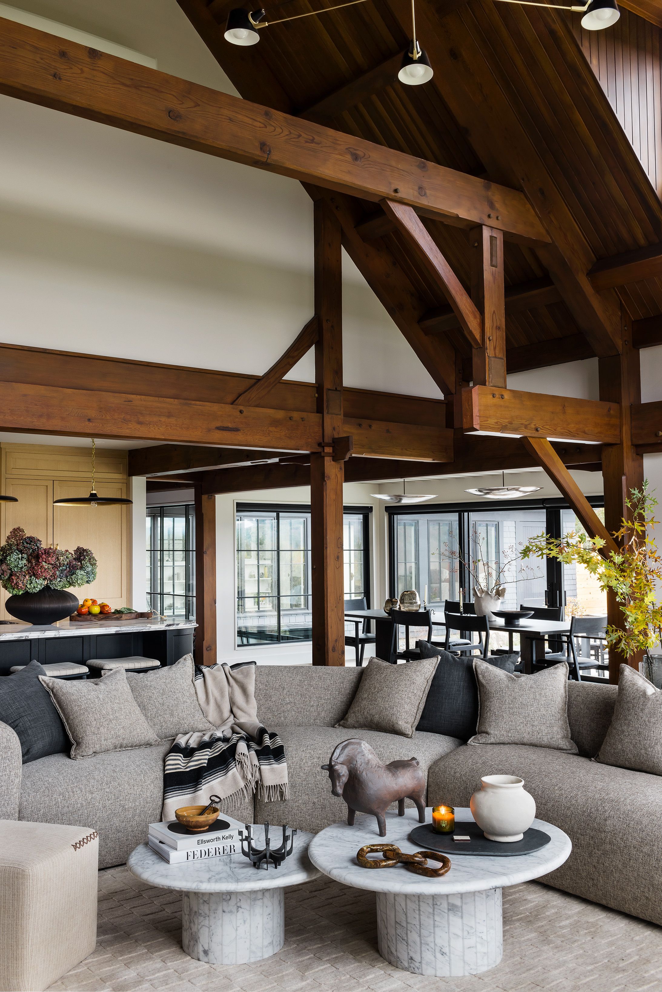A great room with heavy timber trusses and a gray sectional sofa centers on round marble pedestal coffee tables, opening to the kitchen.