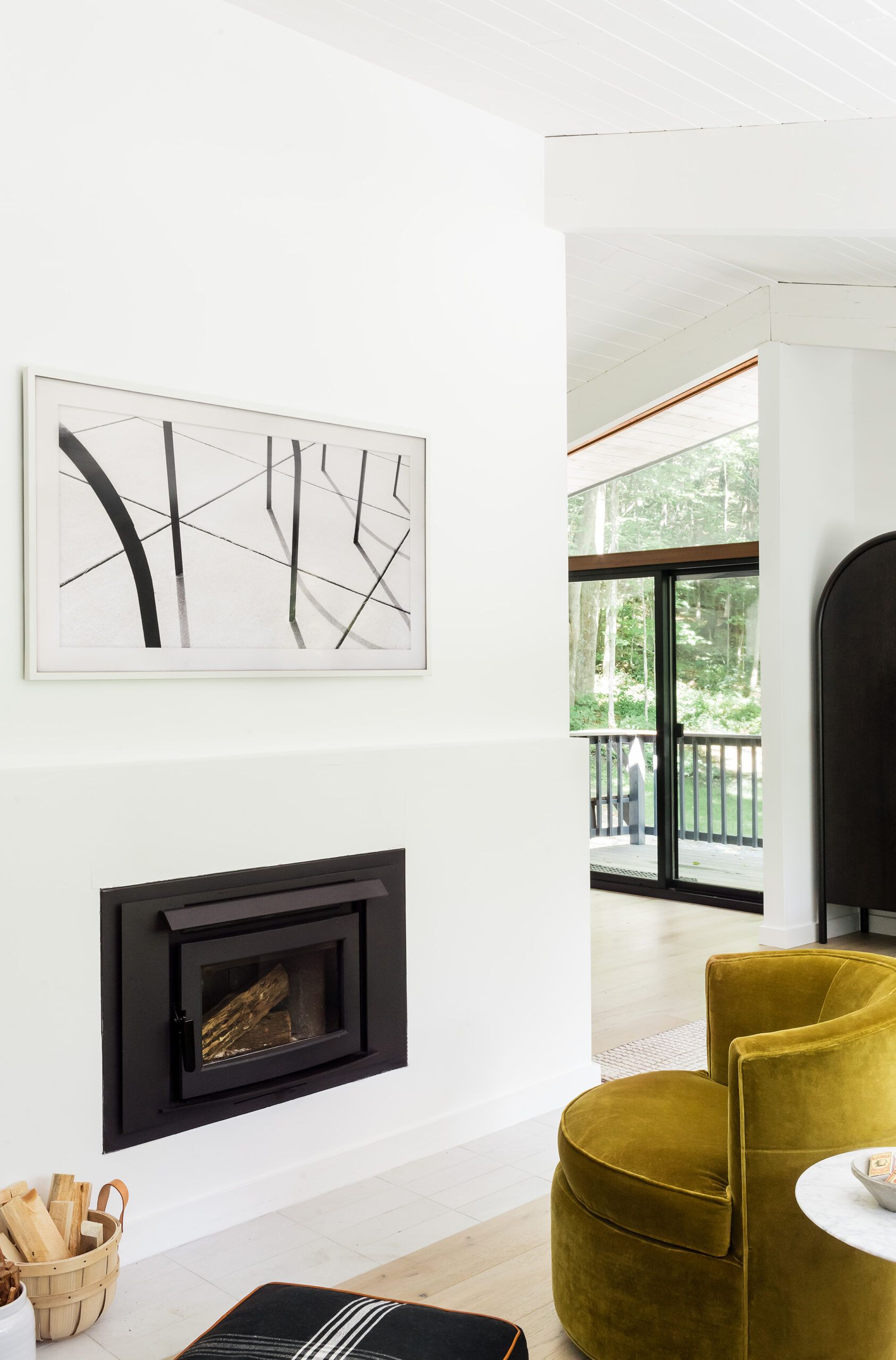 A black fireplace insert with stacked firewood below an abstract black-and-white photograph, beside a mustard velvet chair.