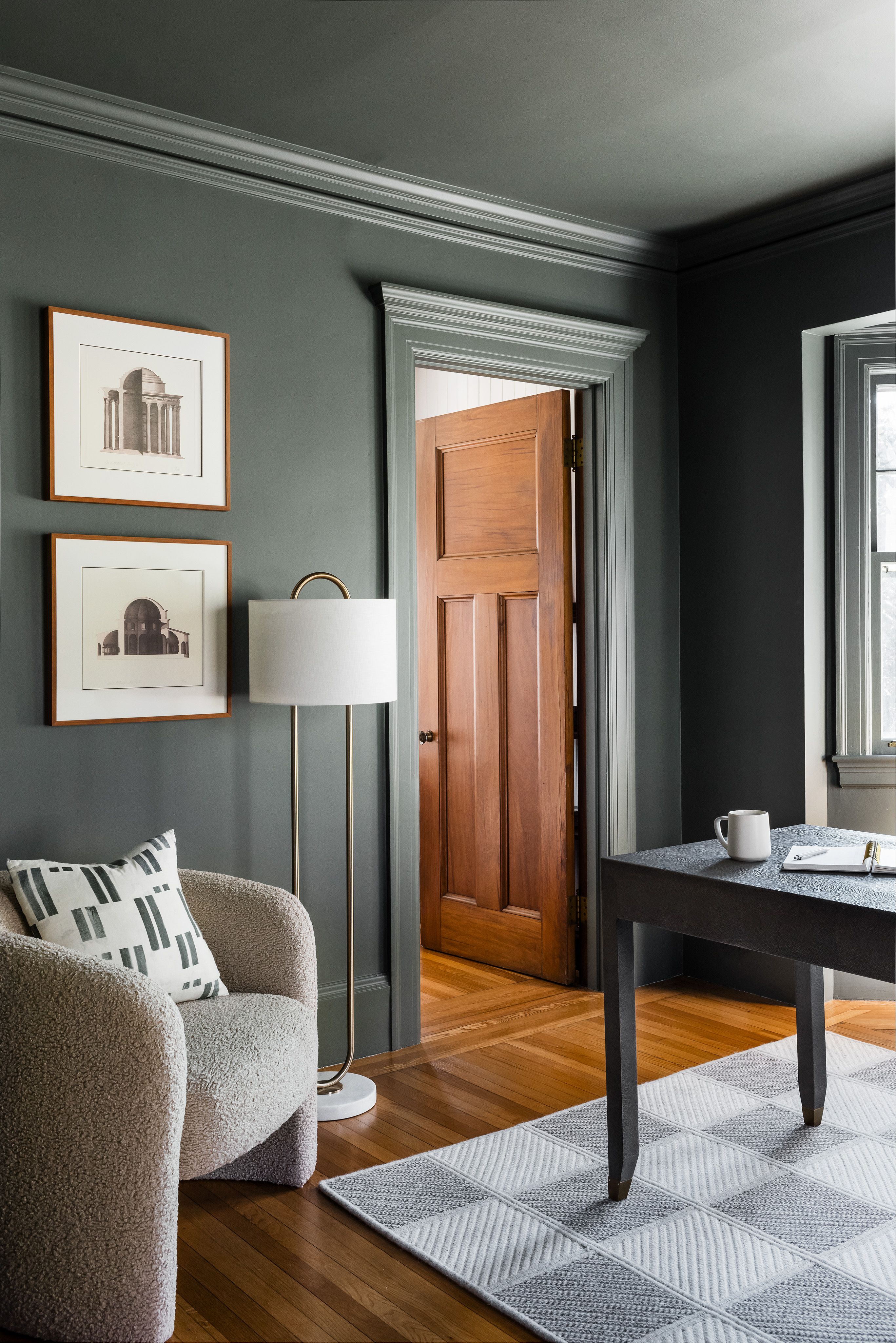 A dark green home office with a gallery of architectural prints, a boucle swivel chair, a brass floor lamp, and a paneled wood door.