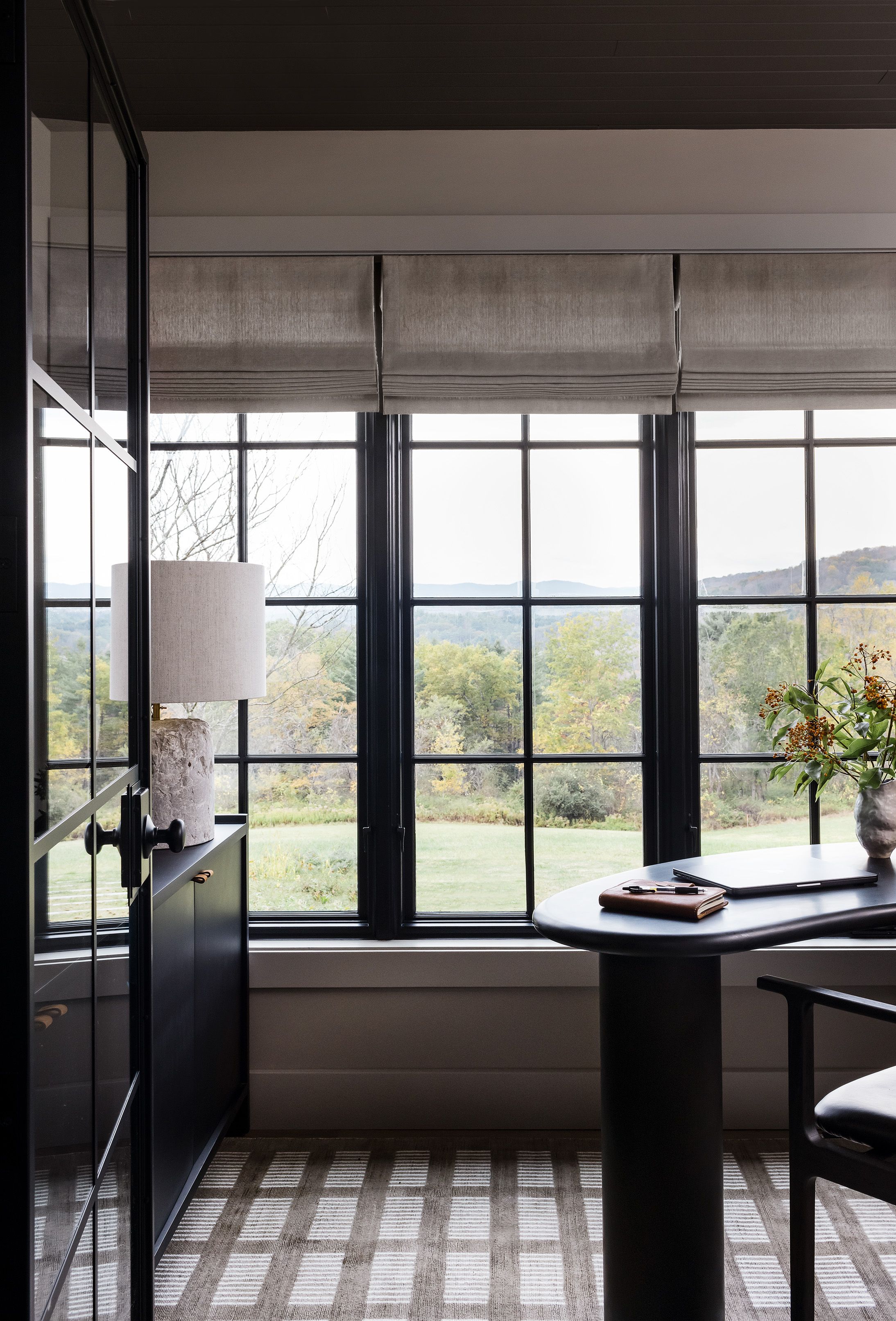 A home office nook has a black pedestal desk and leather chair set before windows overlooking hills, on a plaid wool rug.