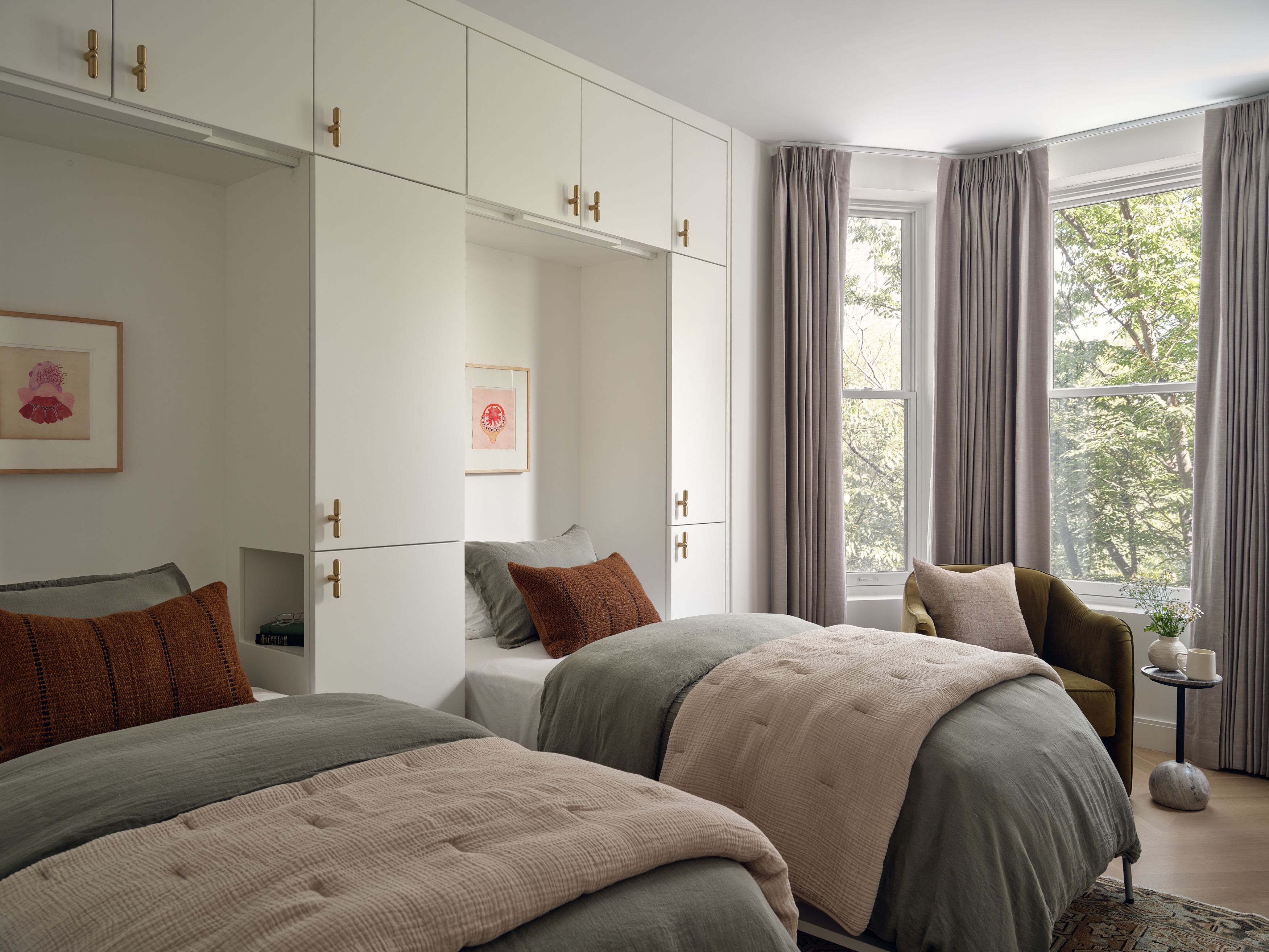 A bedroom with white built-in cabinetry and brass pulls, twin beds dressed in sage green and rust bedding, and framed floral art.