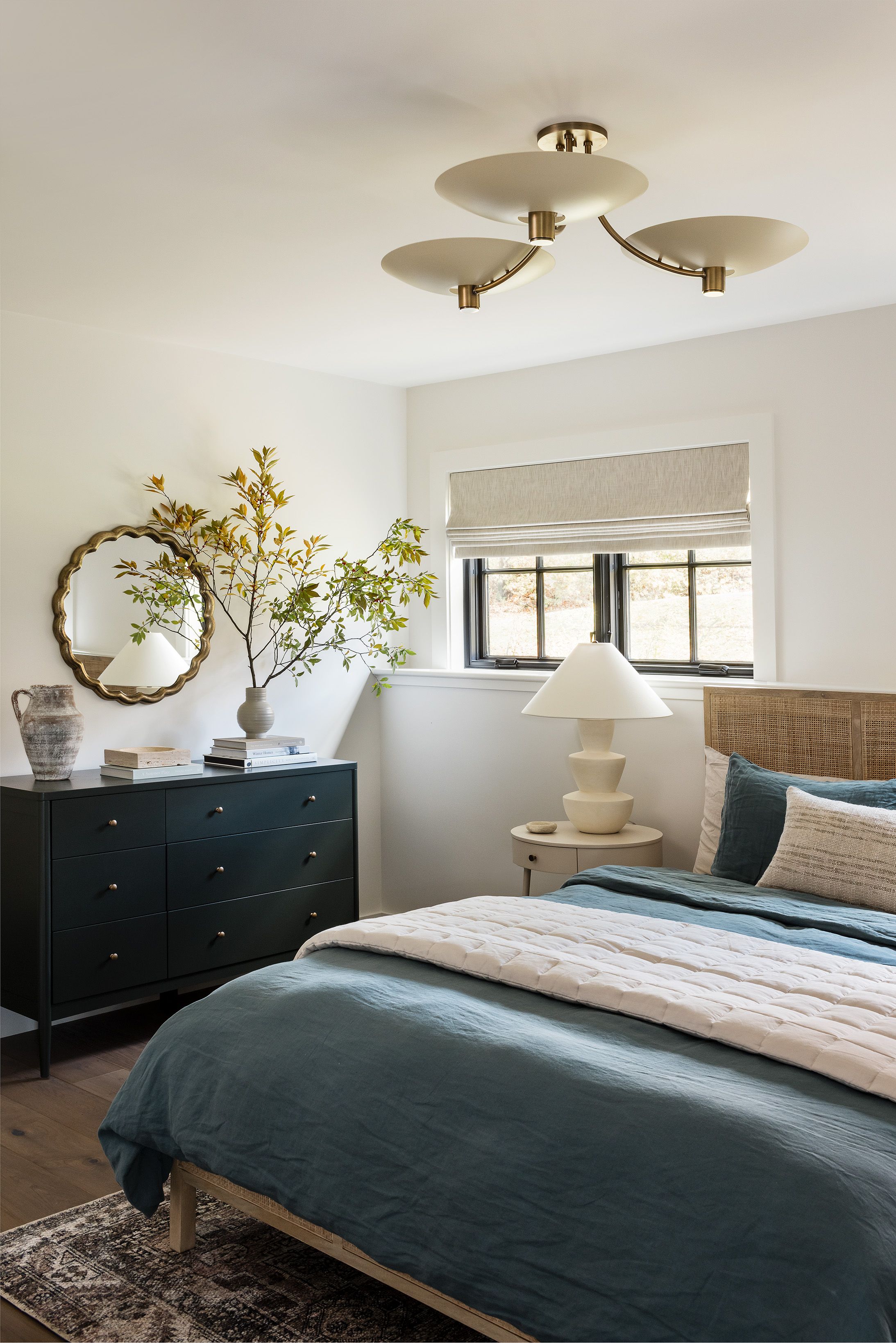 A guest bedroom pairs a rattan headboard and teal linen bedding with a dark green dresser, a scalloped brass mirror, and a three-arm ceiling light.
