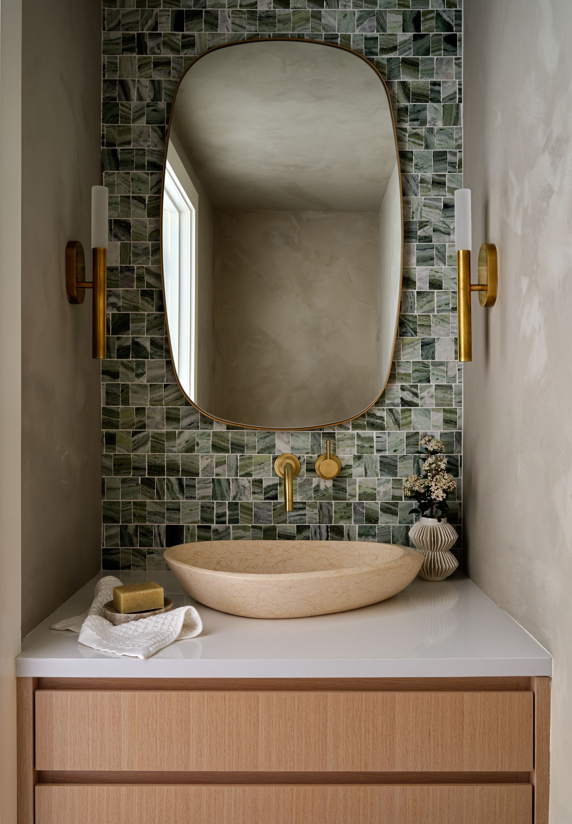 A powder room with floor-to-ceiling green mosaic tile, a brass oval mirror, brass sconces, and a carved stone vessel sink.