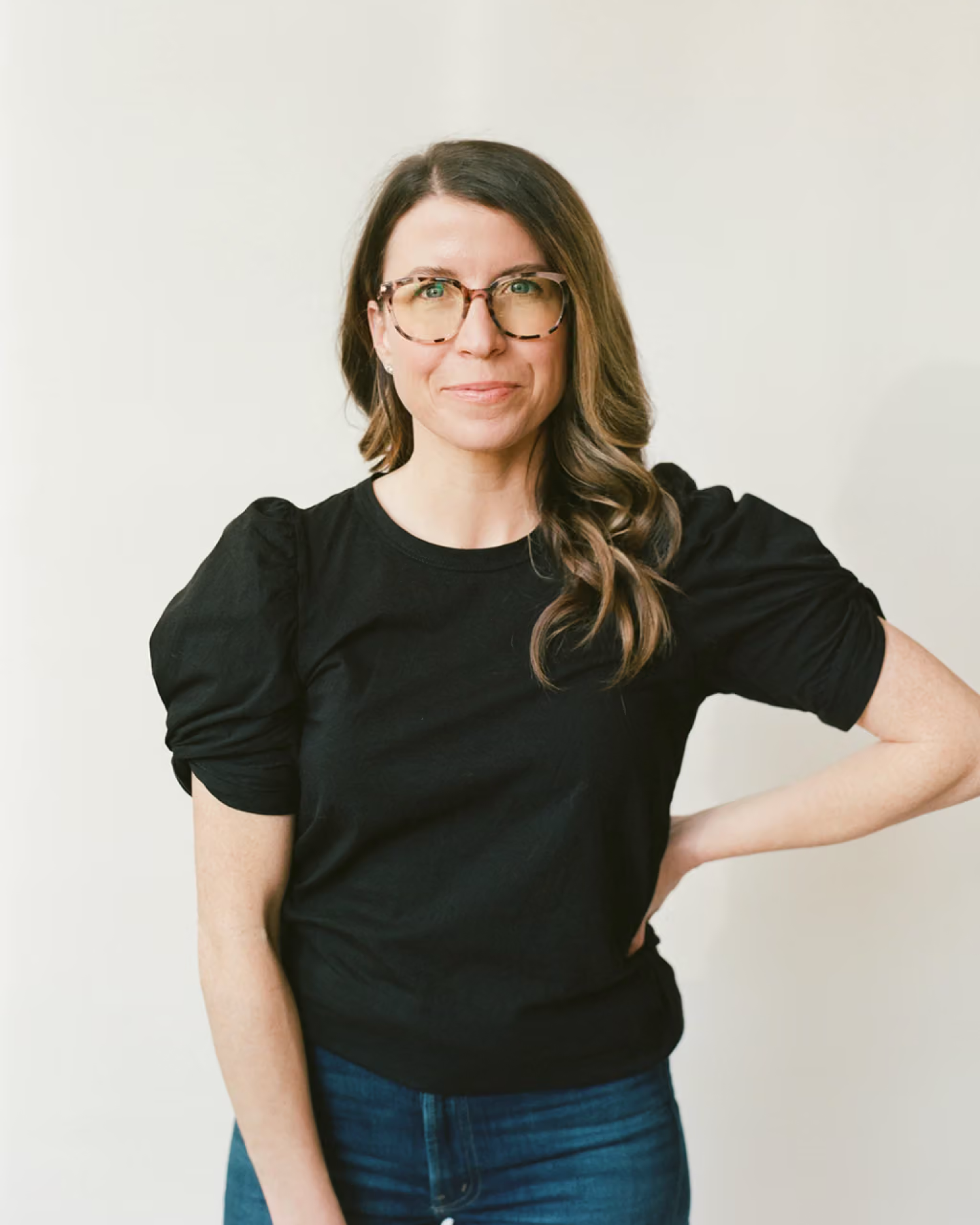 Jess Klein smiles in a black puff-sleeve top and tortoiseshell glasses against a plain white backdrop.