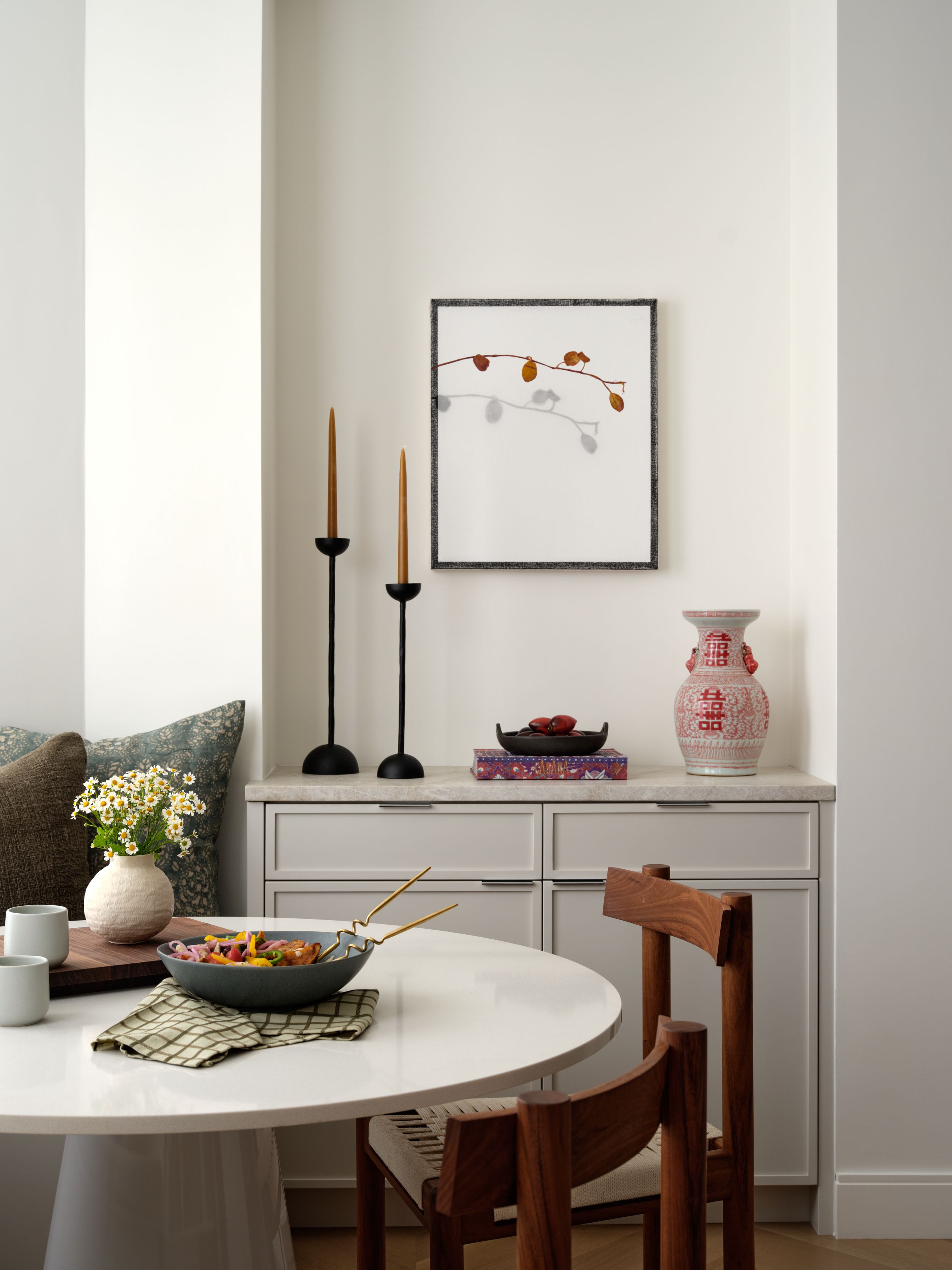 A sideboard vignette with black taper candlesticks, framed branch artwork, a red ginger jar vase, and woven dining chairs.