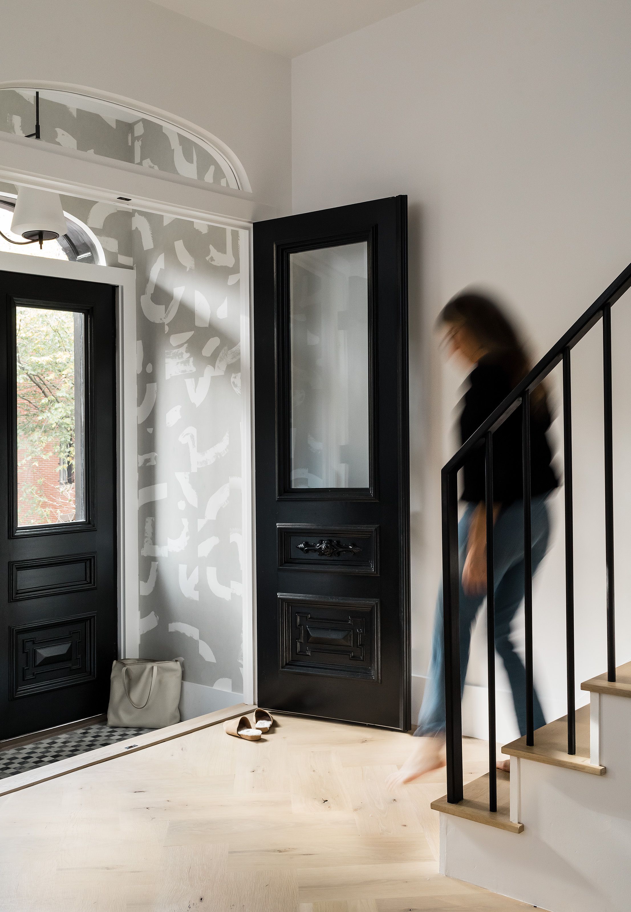 Someone walks up a black-railed staircase past brushstroke-patterned wallpaper and an open black front door with checkerboard tile.