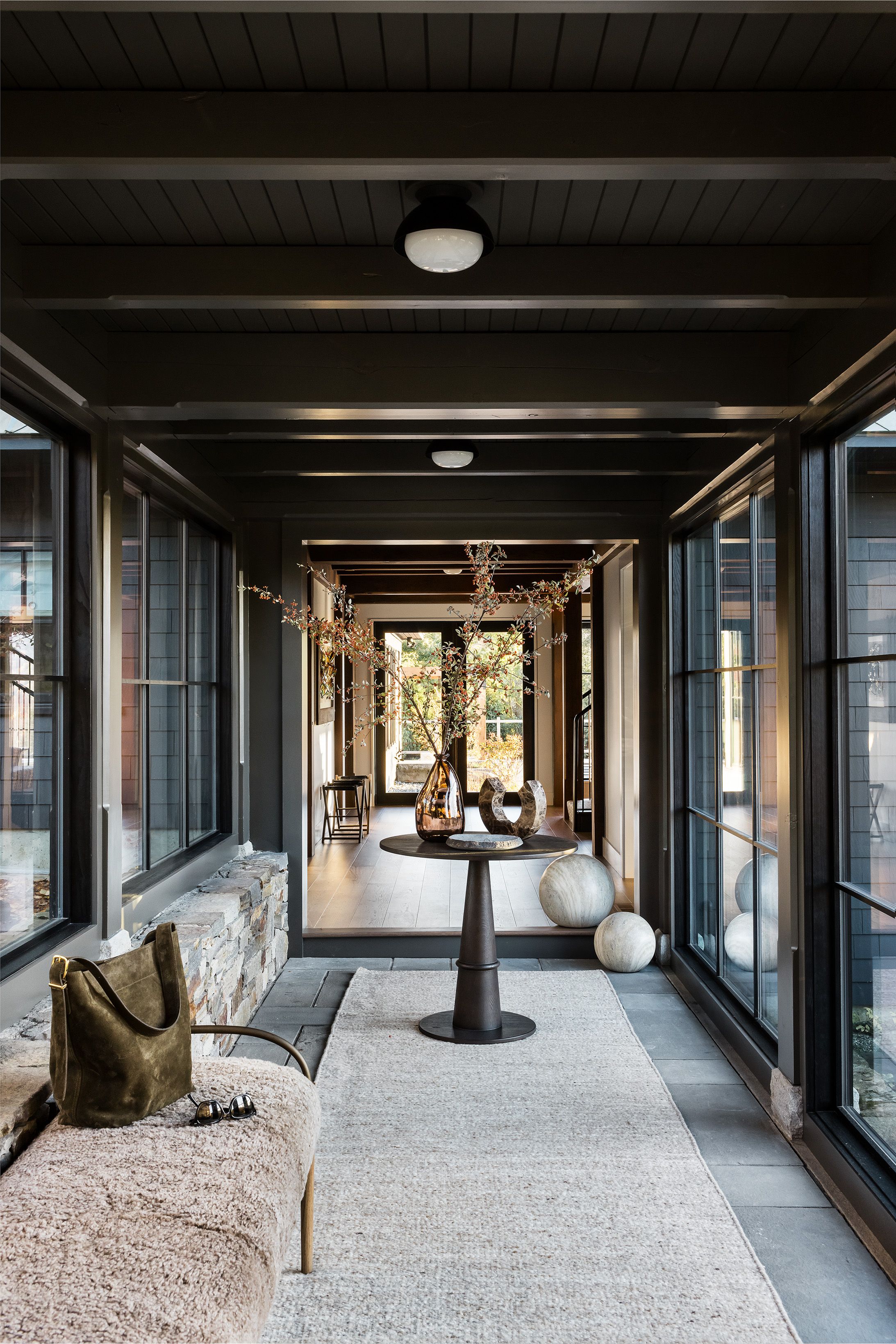 A glass-walled breezeway with a stone half-wall leads past a round pedestal table and a shearling bench toward the home's entry.