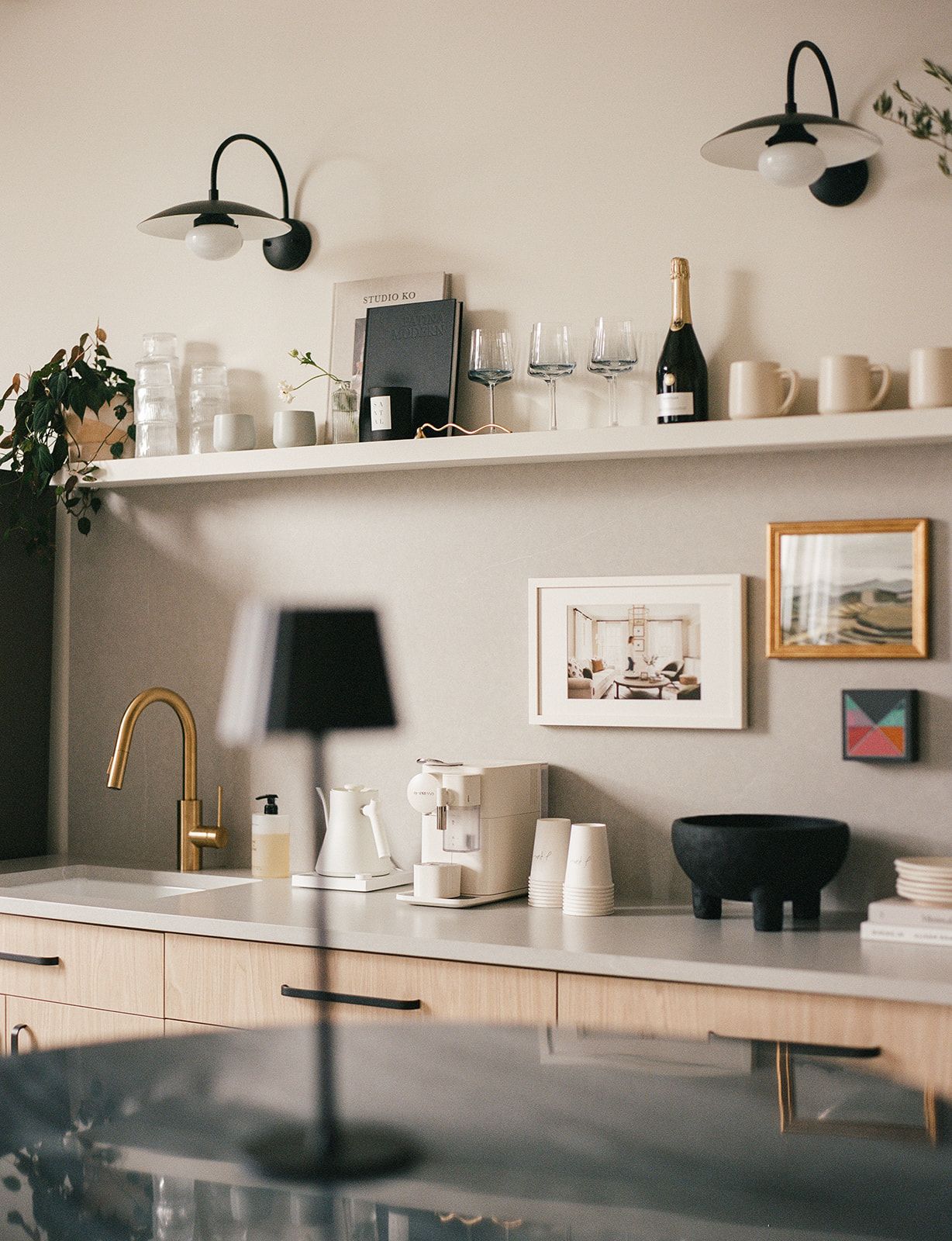 A studio bar counter with a brass faucet, a blurred black table lamp, an open shelf of wine glasses and champagne, and a gallery wall of art.