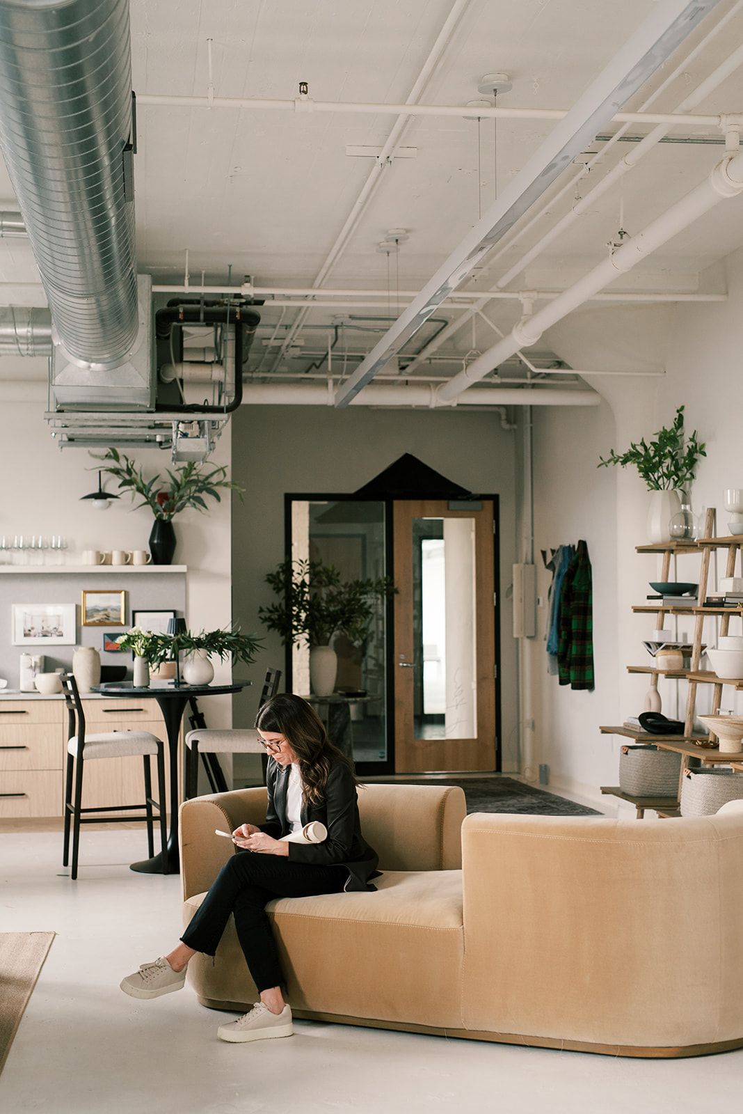 Someone relaxes on a camel velvet sofa in an open kitchen with pale wood cabinetry, bar stools, and ladder shelving.