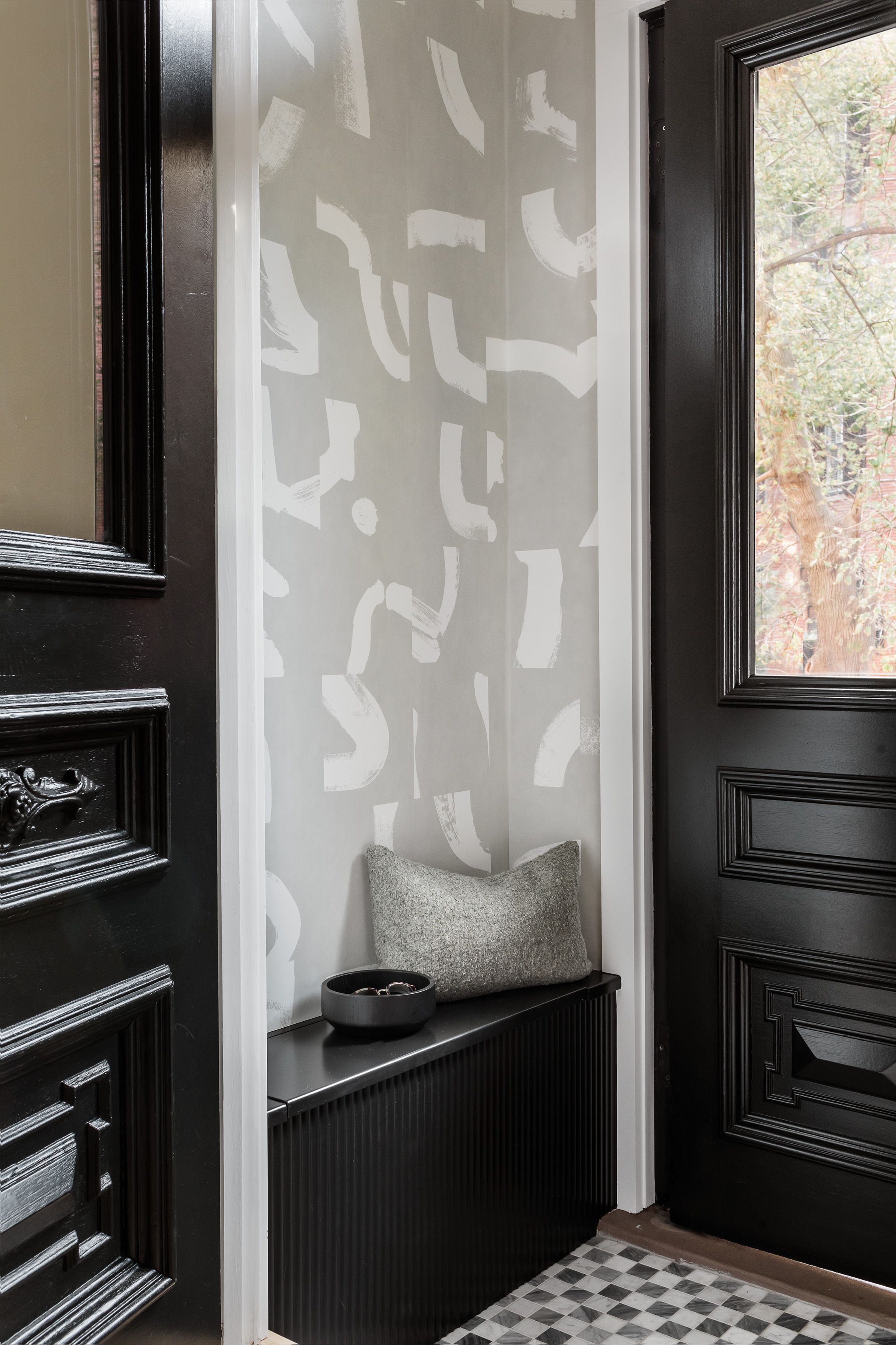 Entry with a black paneled front door, gray brushstroke-patterned wallpaper, a black bench, and light oak herringbone flooring.
